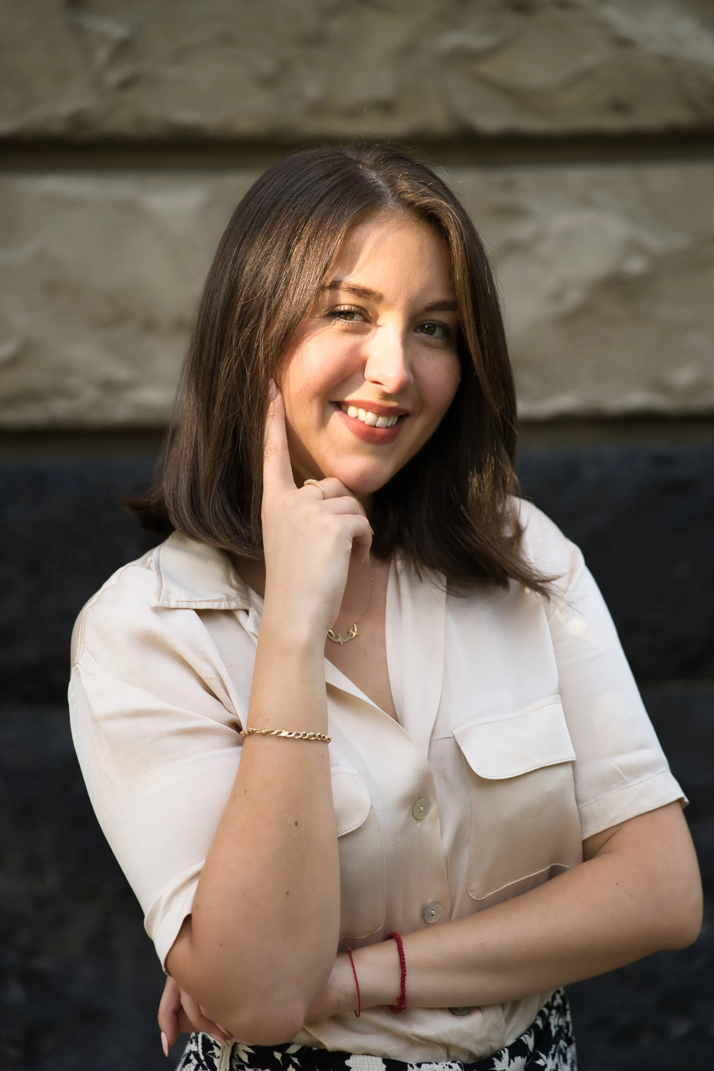 A portrait of a young woman with shoulder-length brown hair, smiling, wearing a beige short-sleeved blouse, gold jewelry, and a red bracelet, standing in front of a stone or concrete wall.