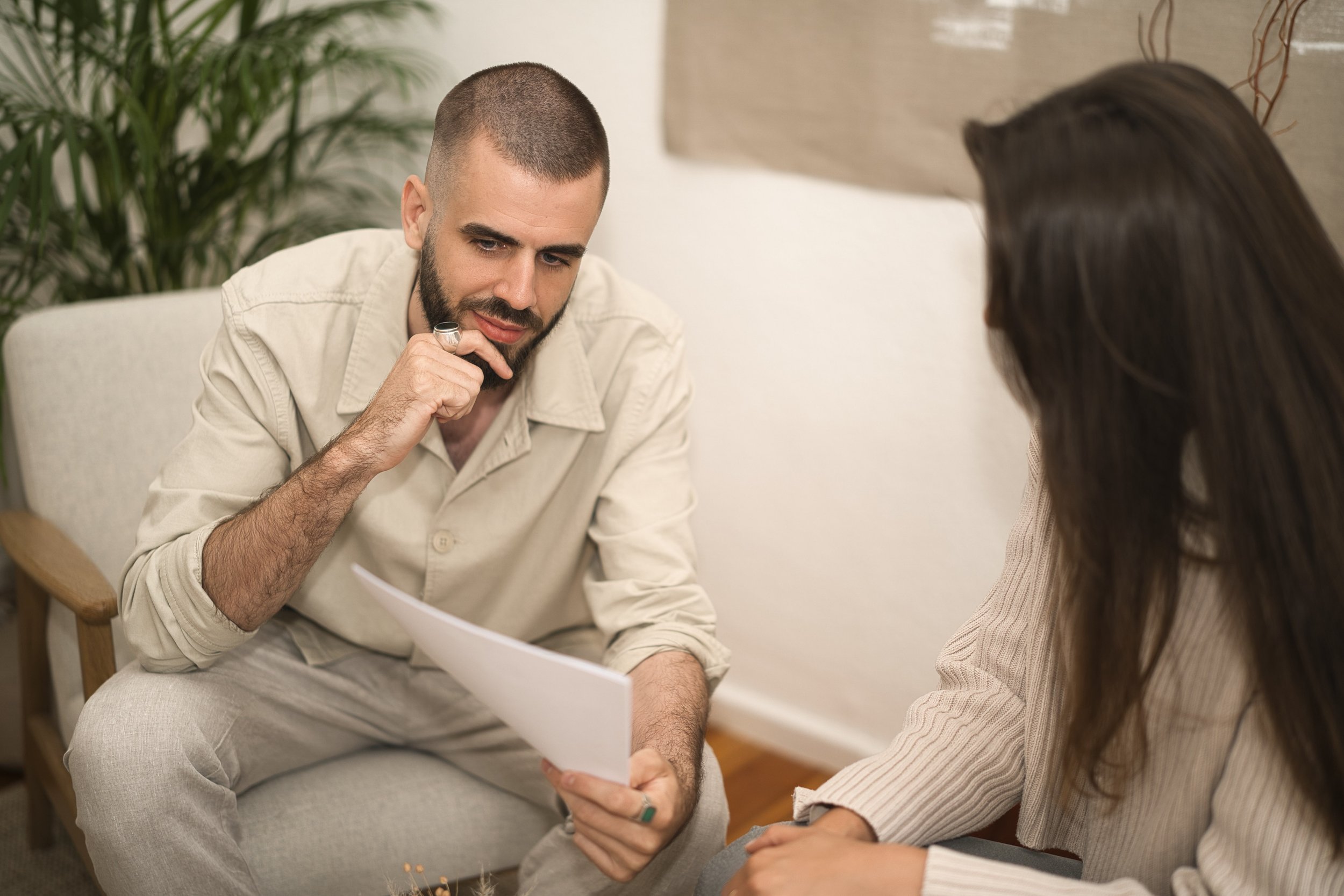 A business man with a beard and short hair looks at a business woman with brown hair during a conversation in a cozy room, with plants and artwork in the background.
