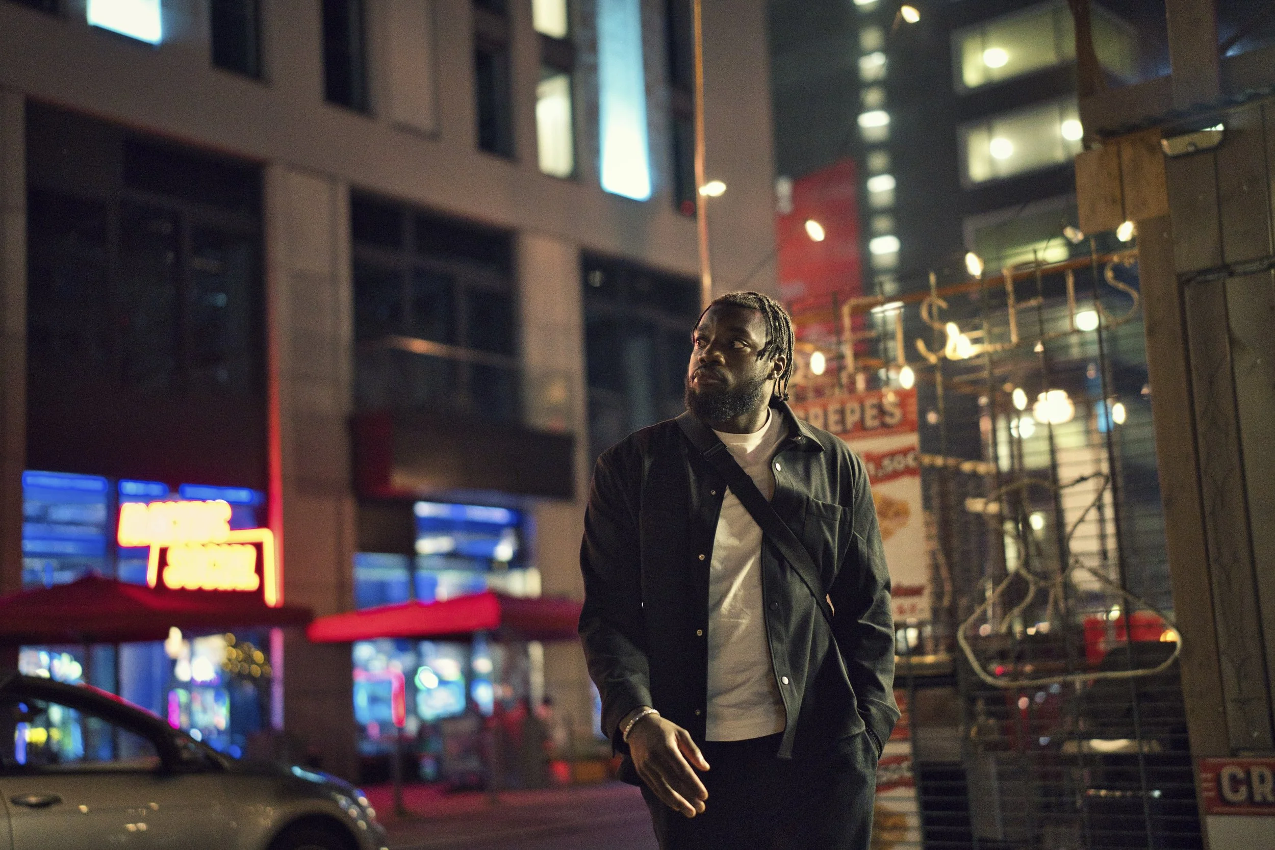 A portrait of a man with braided hair, wearing a black jacket and white t-shirt, walking on a city street at night with neon lights, buildings, and a sign in the background.