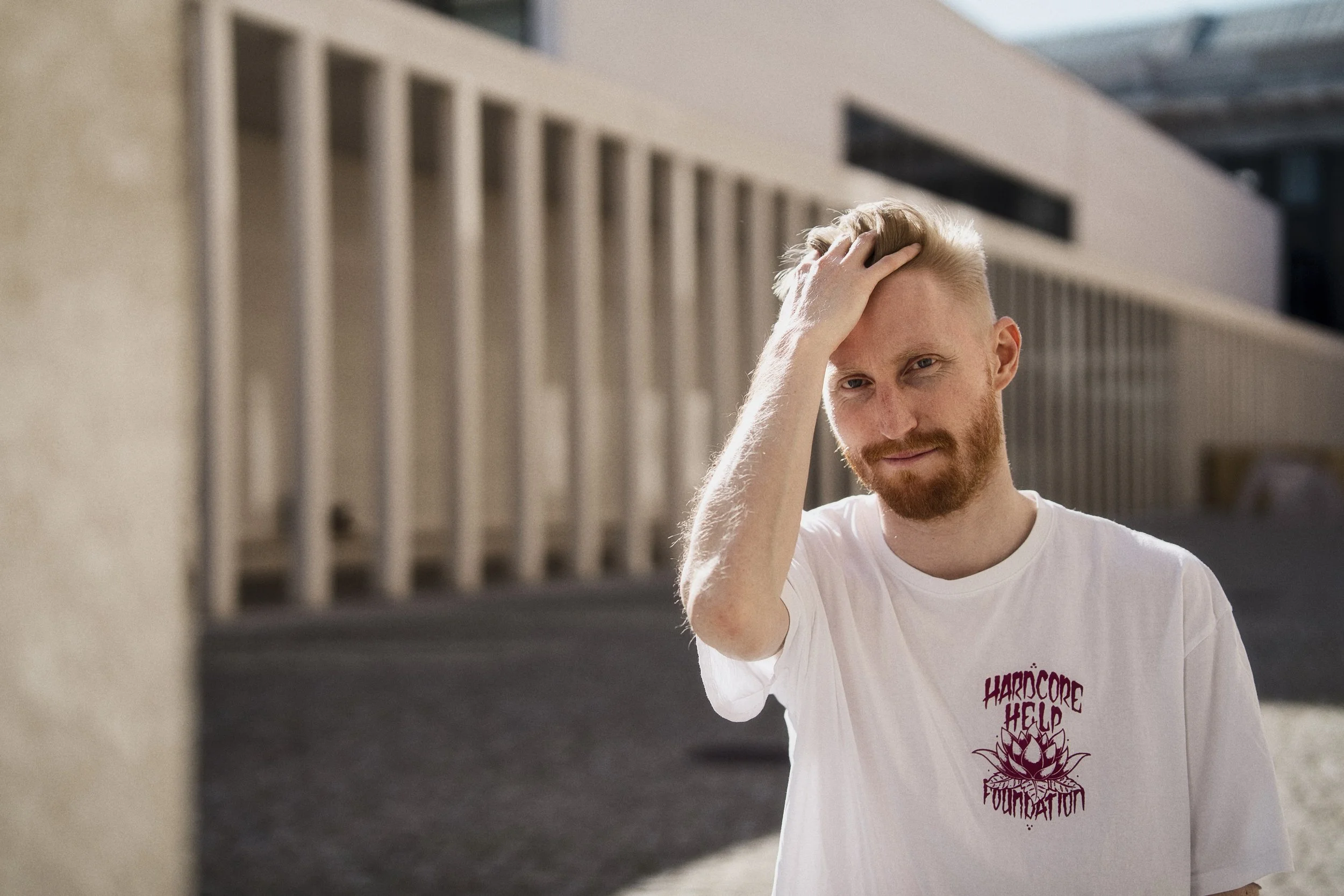 A portrait of a man with red hair and a beard touches his head with one hand, standing outdoors in sunlight. He wears a white t-shirt with a purple design on it.