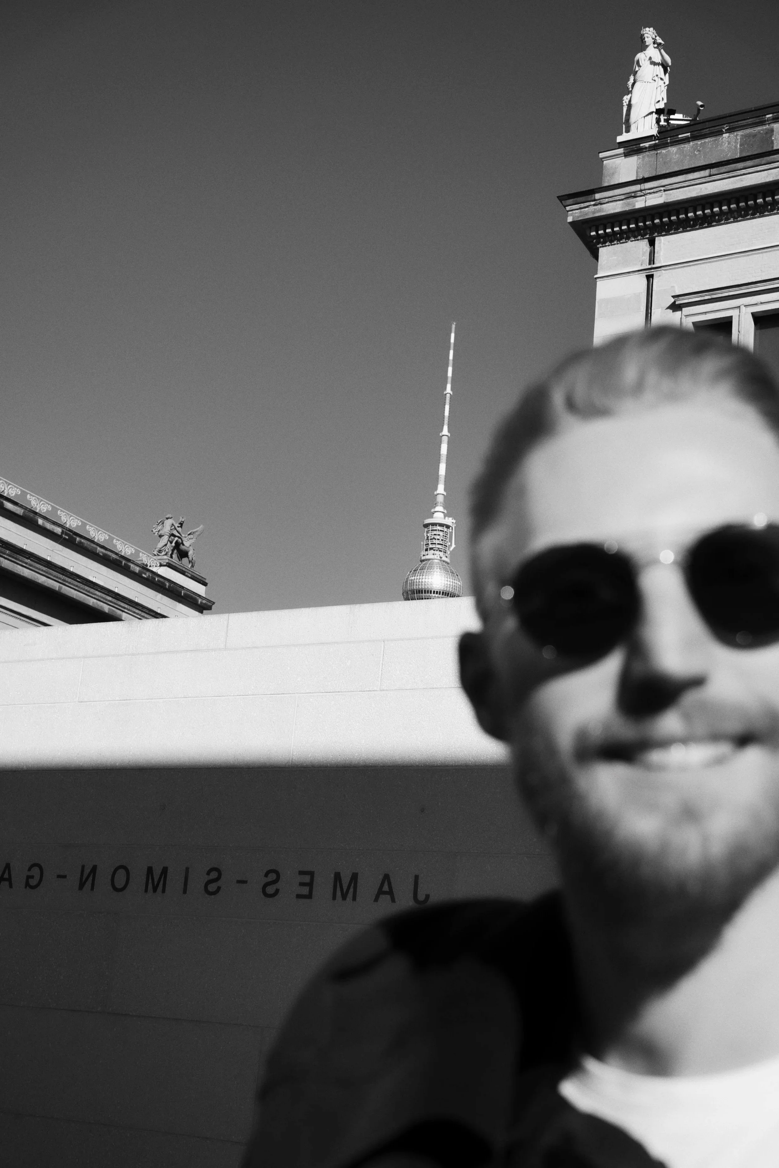 Black and white portrait of of a person wearing sunglasses, with a cityscape featuring a tall tower and classical statues in the background.