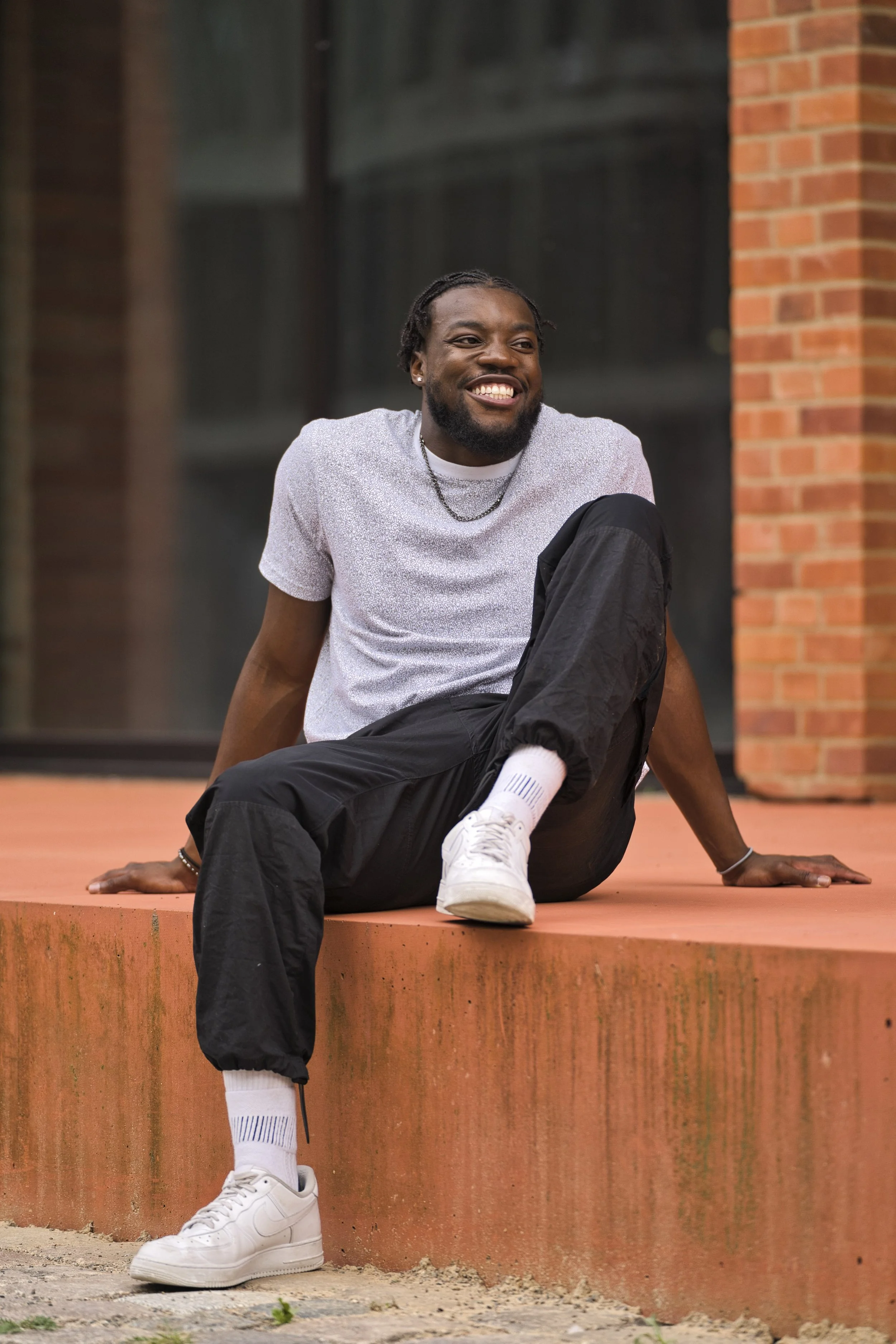 A portrait of a smiling man with dreadlocks and a beard, wearing a white t-shirt, black pants, white sneakers, and white socks, sitting on an orange ledge outdoors.