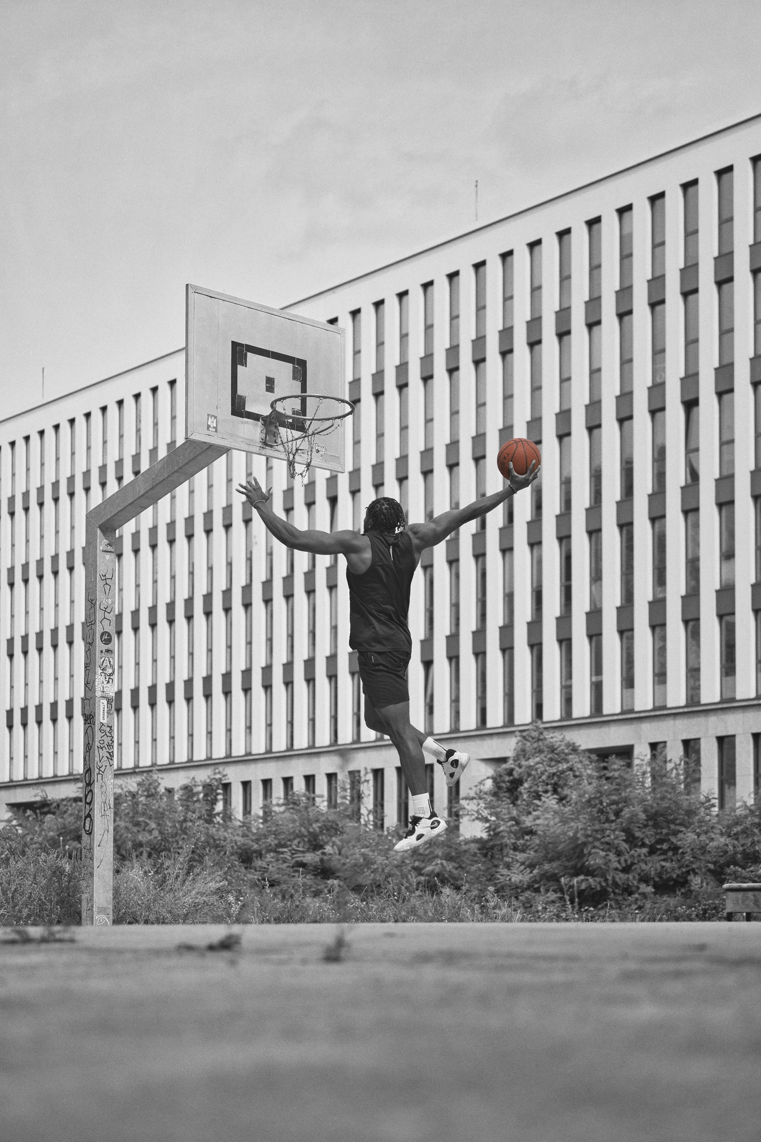 A portrait of a person playing basketball outdoors, jumping to make a shot, with a tall, modern building in the background, in black and white.