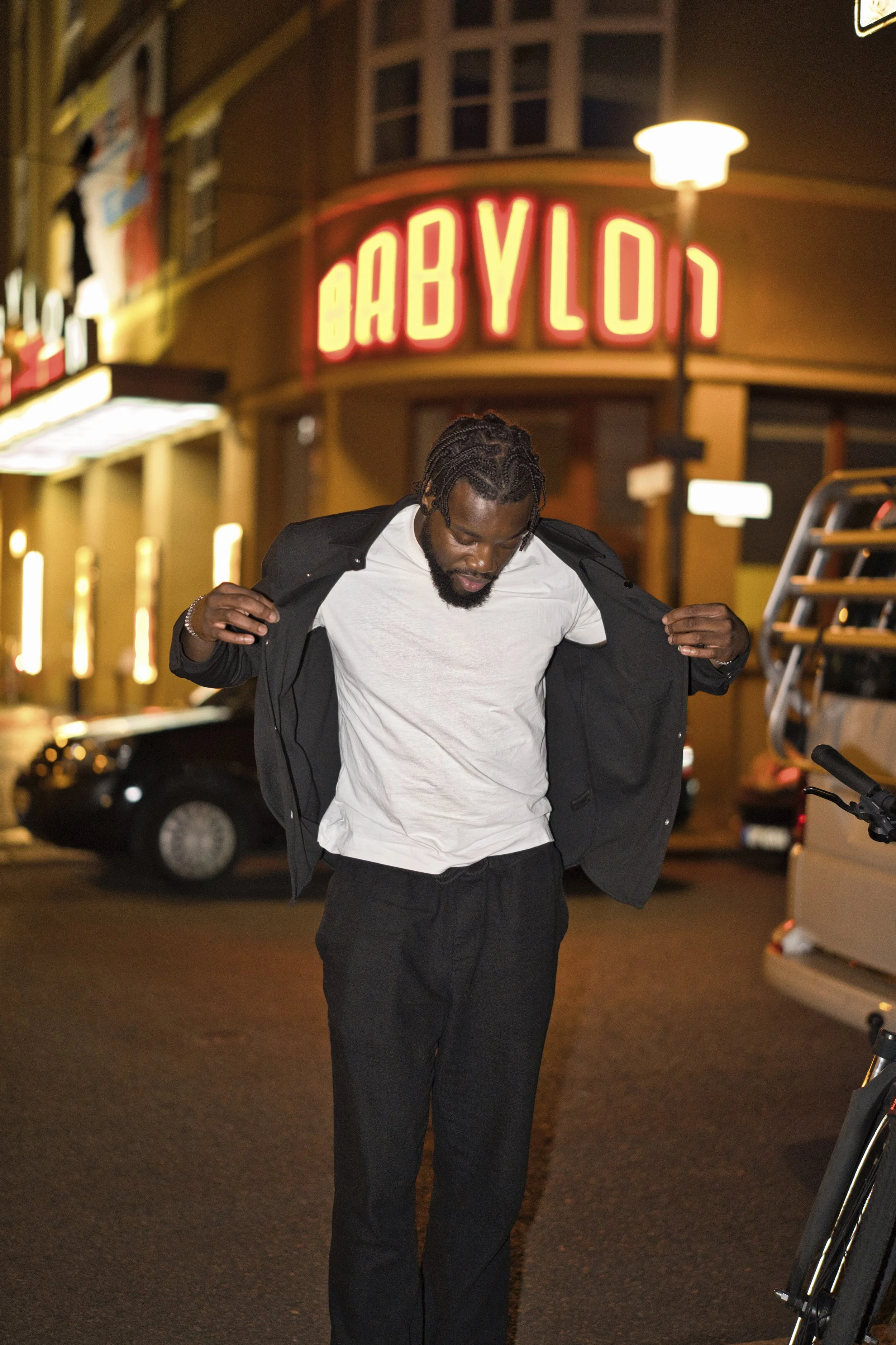 A portrait of a man with braided hair and a beard is putting on a black jacket while standing on a city street at night. In the background, there is a neon sign that reads 'BABYLON' and some parked cars.