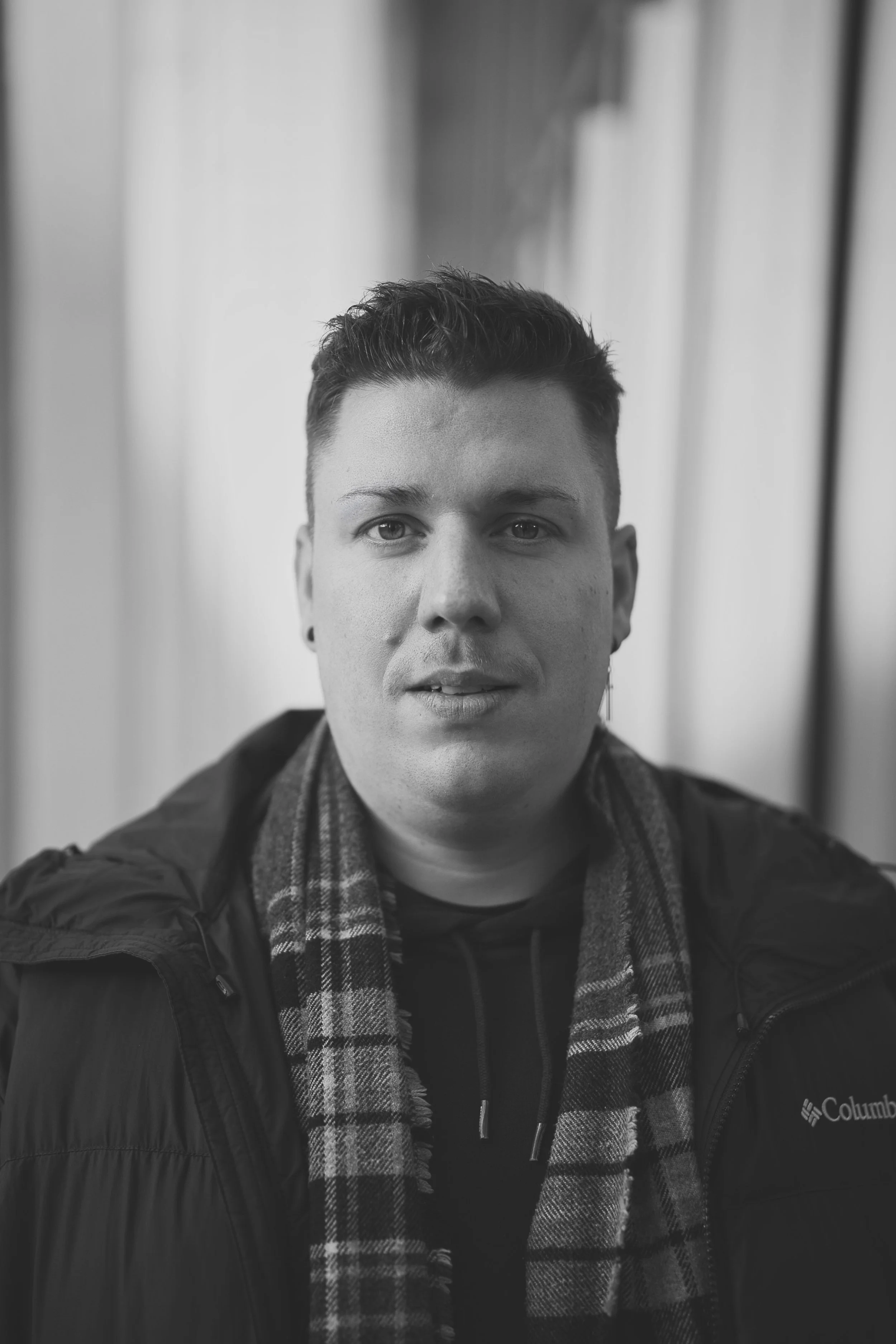 A portrait of a young man with short dark hair and earrings taking a selfie in black and white. He wears a dark jacket and a plaid scarf, with a neutral facial expression. The background is blurred.