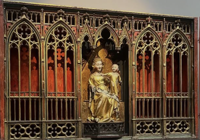 Gold-colored statue of a woman holding a child, framed by ornate gothic-style metal gate with pointed arches and floral patterns, set against a red wall in a church or cathedral.