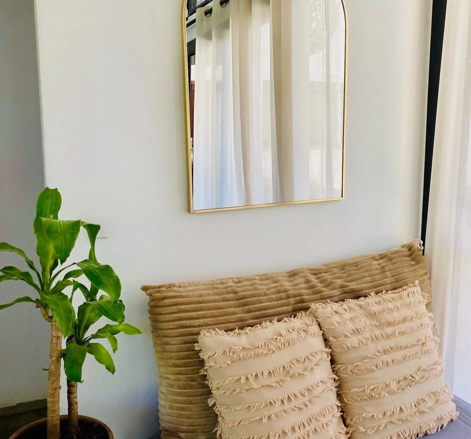 A beige sofa with ruffled cushions in front of a white wall, with a rectangular mirror and a potted green plant nearby.