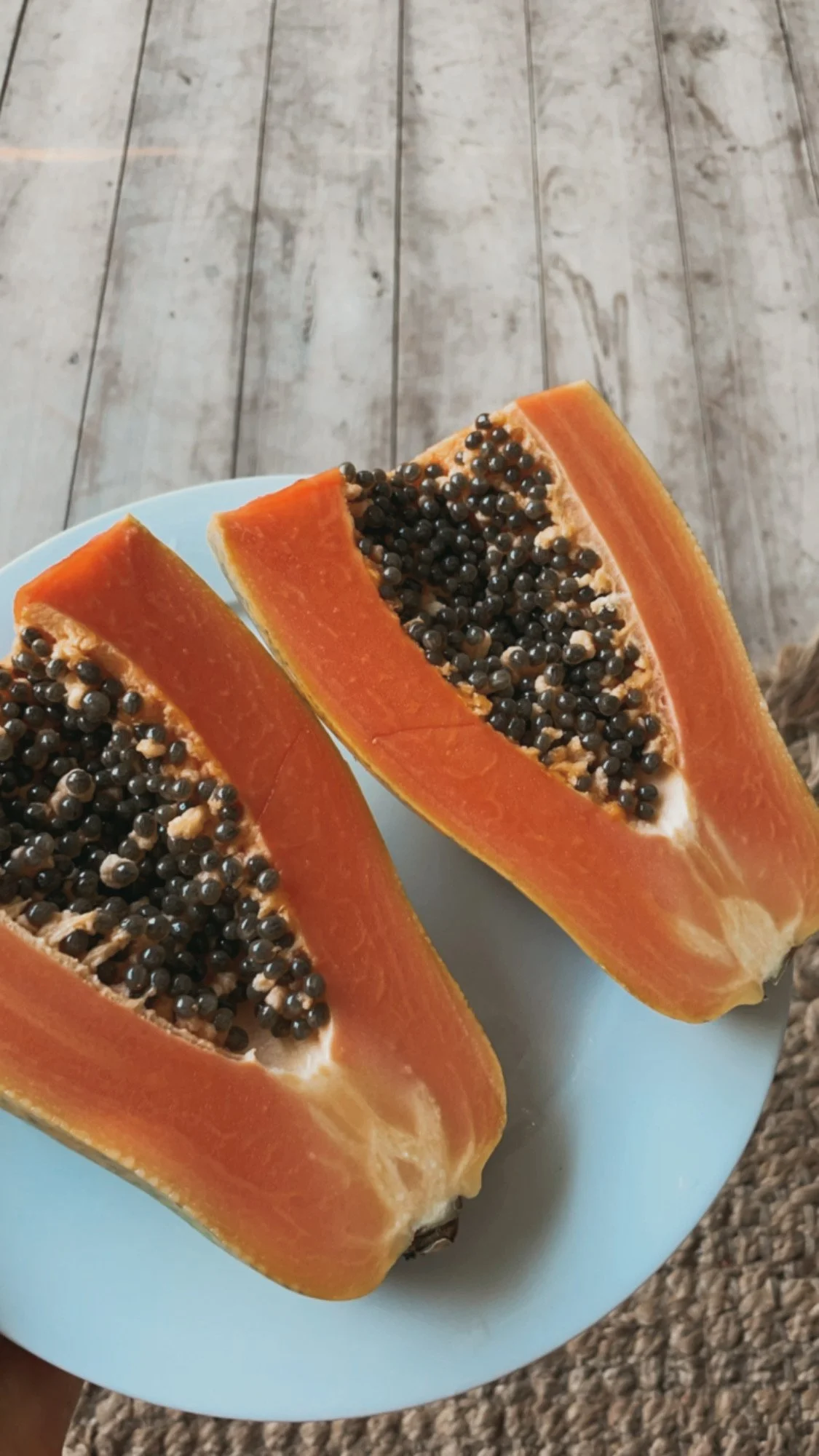 Two halves of a papaya fruit on a white plate, showing orange flesh filled with black seeds, on a wooden surface.