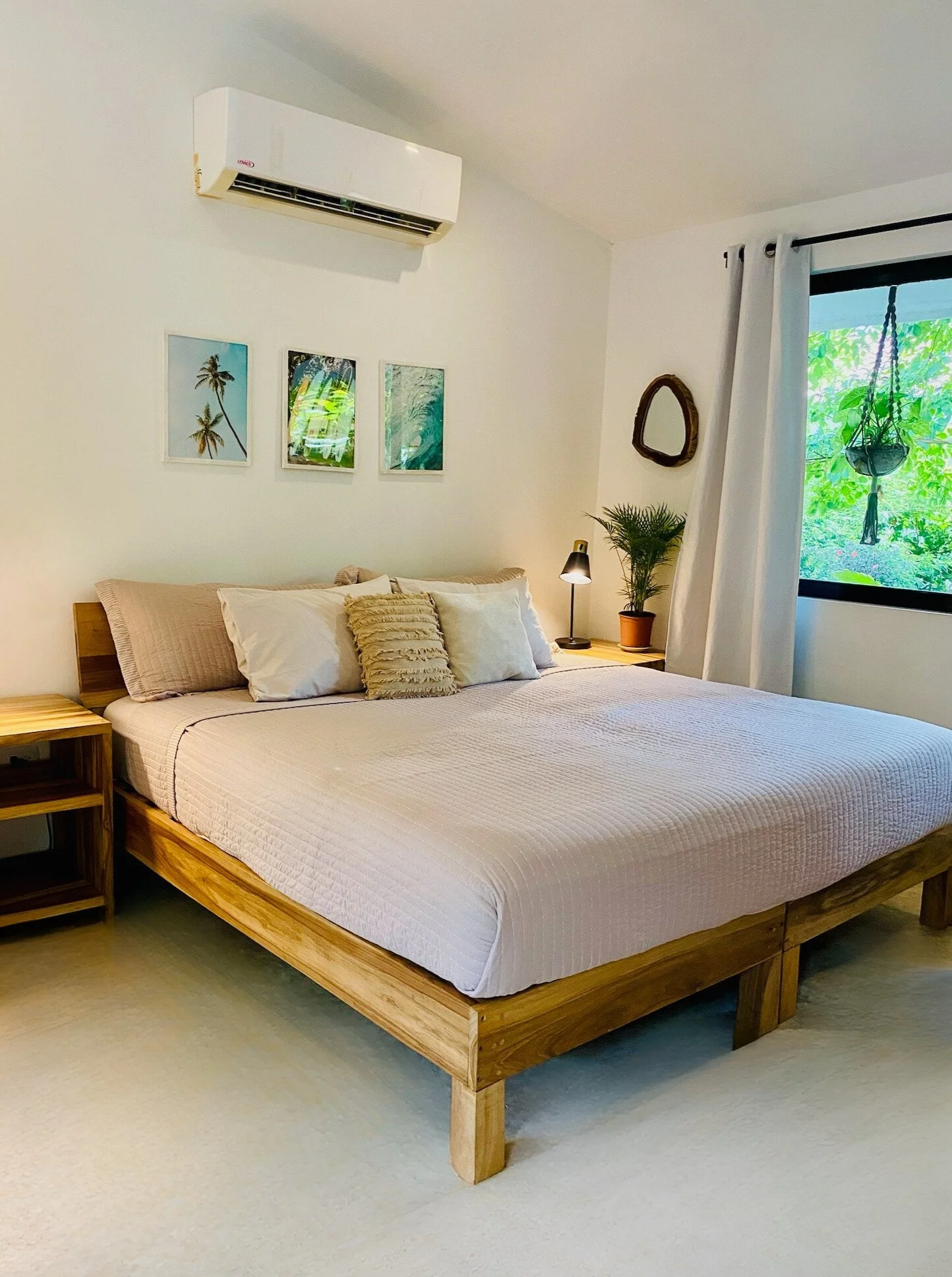 A bedroom with a wooden bed frame, white bedding, and multiple pillows, with a nightstand and lamp on the side. There's a window with white curtains, a small plant, and three framed pictures of tropical scenes on the wall. An air conditioner is mounted above the bed.