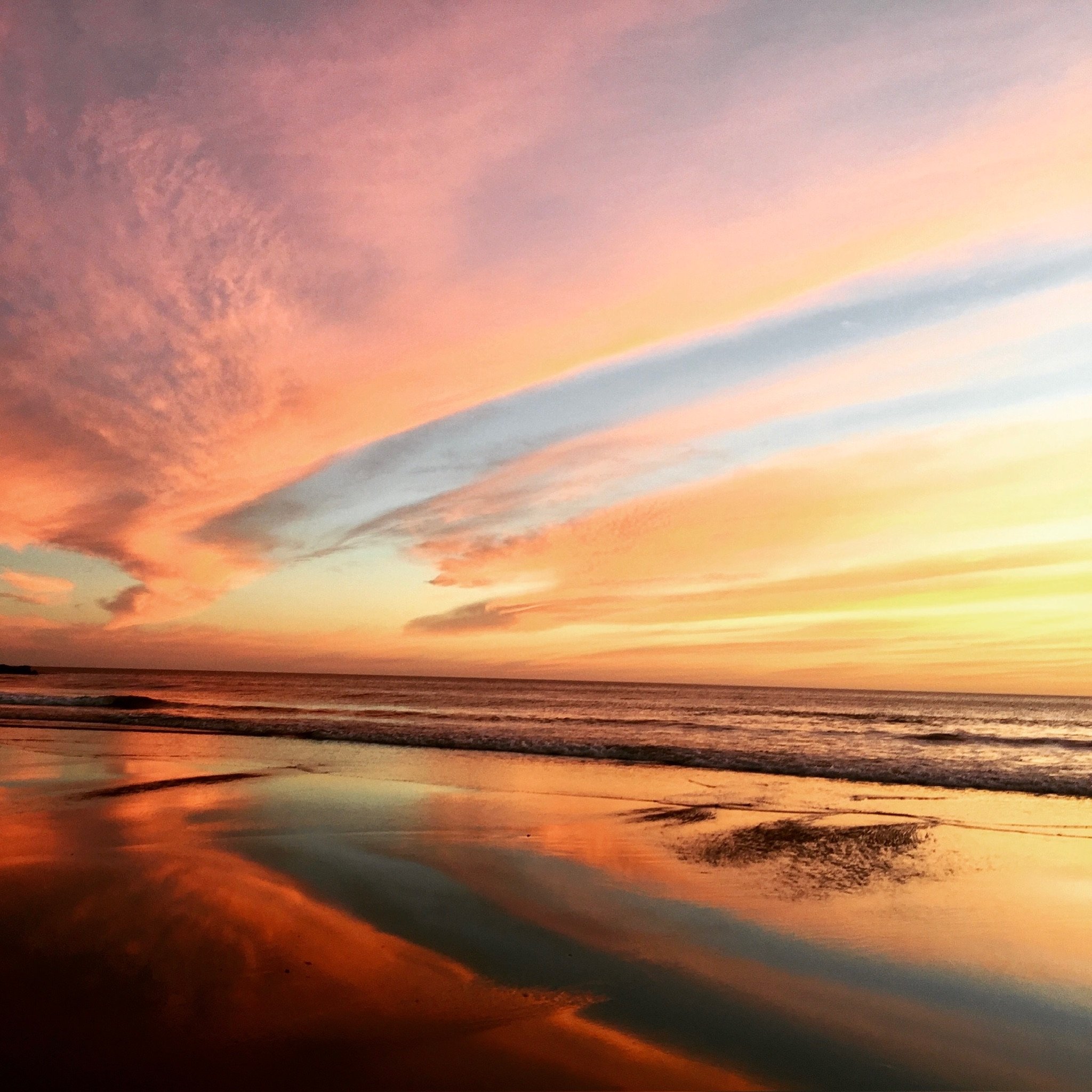 Sunset over the ocean with colorful pink, orange, and blue clouds reflected on the wet sand of the beach.