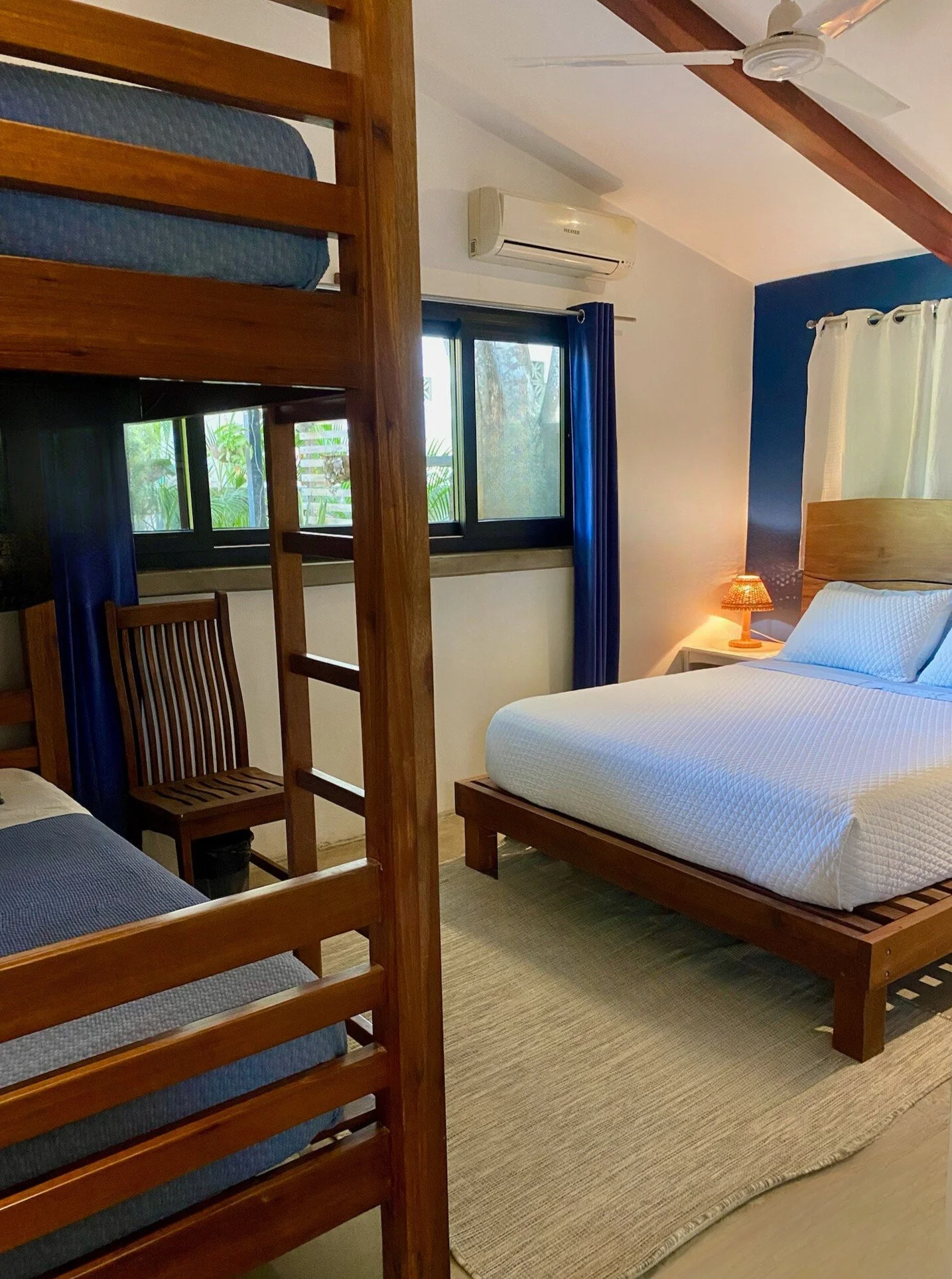 A bedroom with a double bed with white bedding, a wooden headboard, and a small bedside lamp turned on. There is a window with blue curtains behind the bed, next to a wall-mounted air conditioning unit. To the left, there are two sets of wooden bunk beds and a wooden chair.