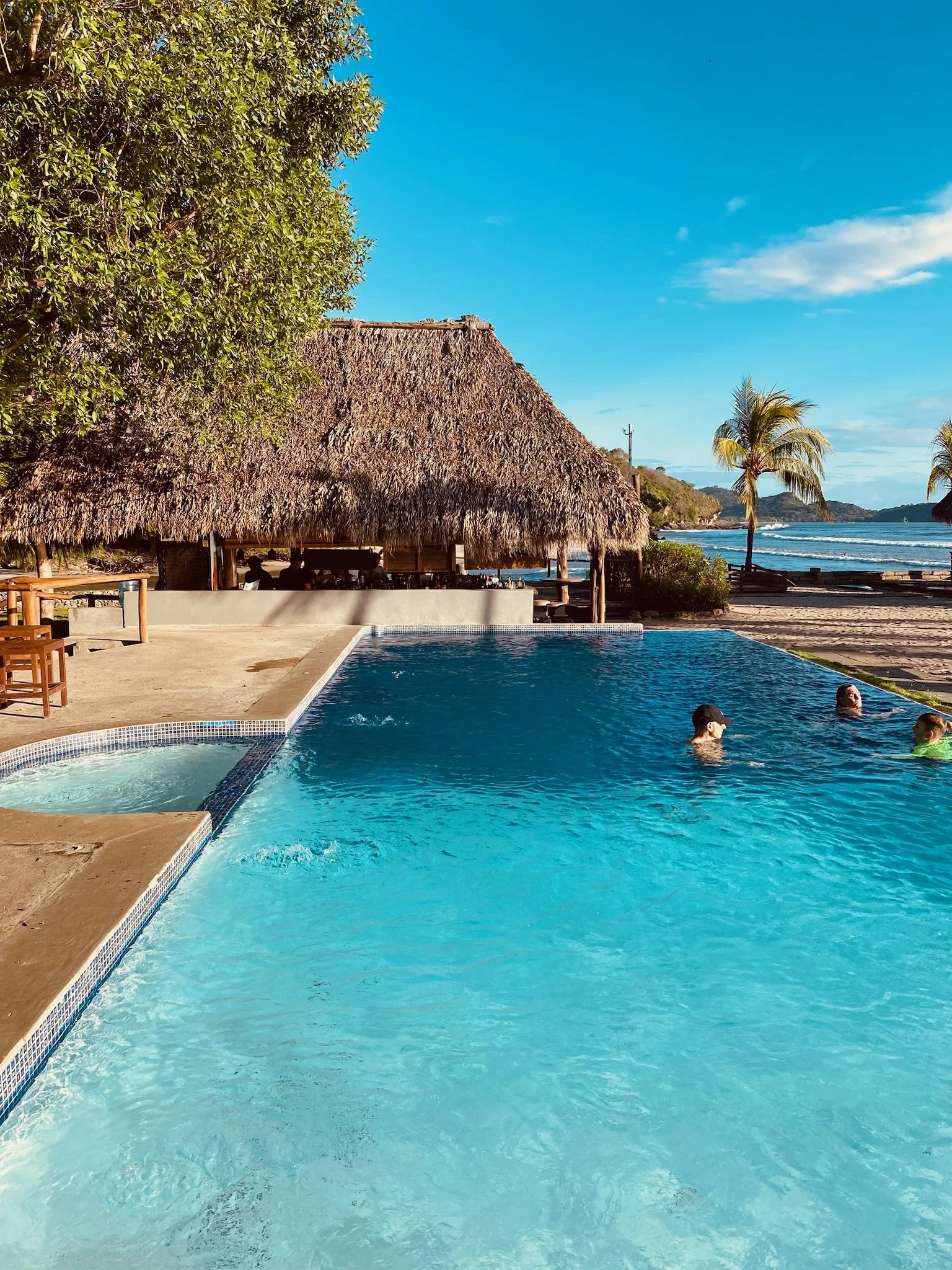 People swimming in a pool near a beach with palm trees and a thatched-roof hut, under a blue sky.