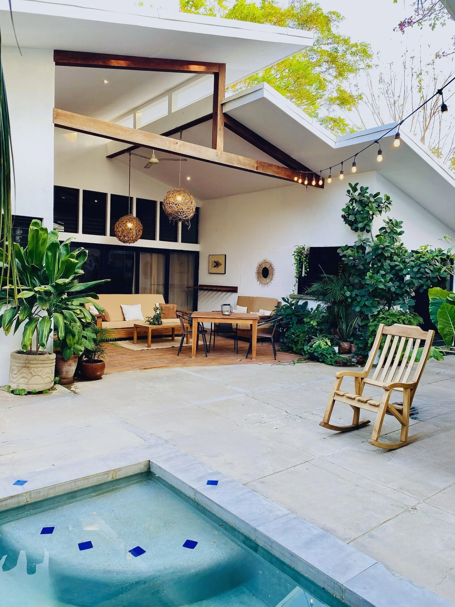 Modern backyard patio with potted plants, outdoor seating, rockers, and string lights, adjacent to a small pool.