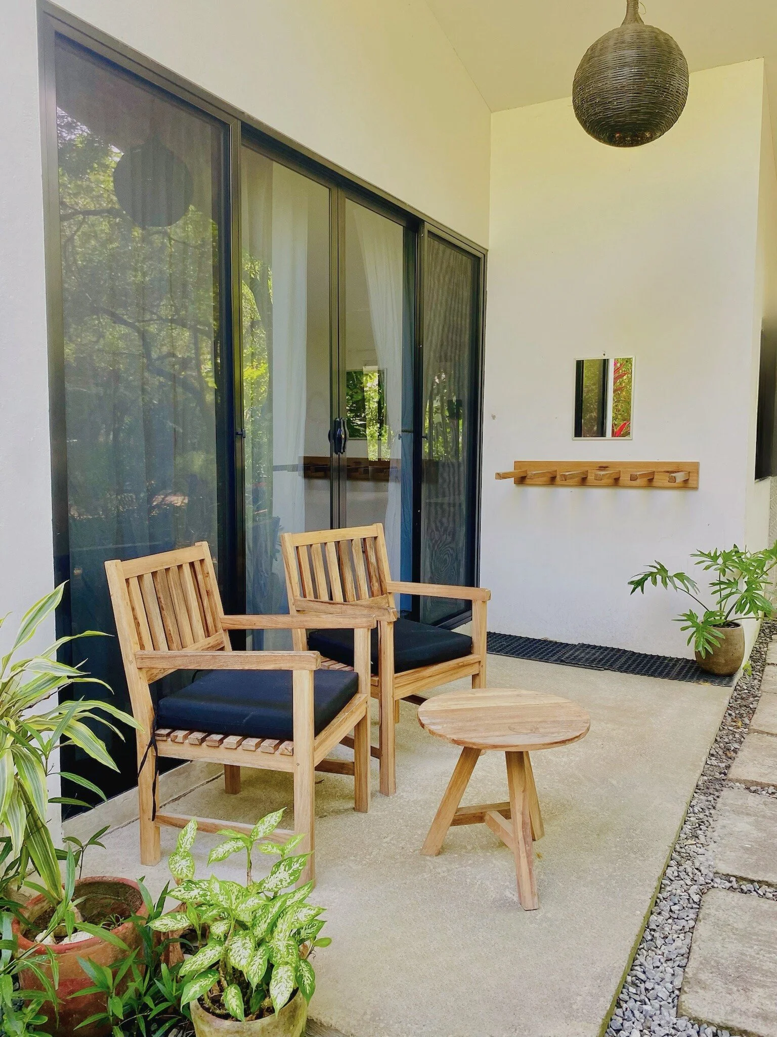 Outdoor patio with two wooden chairs with black cushions, a small wooden table, potted plants, a coat rack, a mirror, a black rug, and a black paper lantern