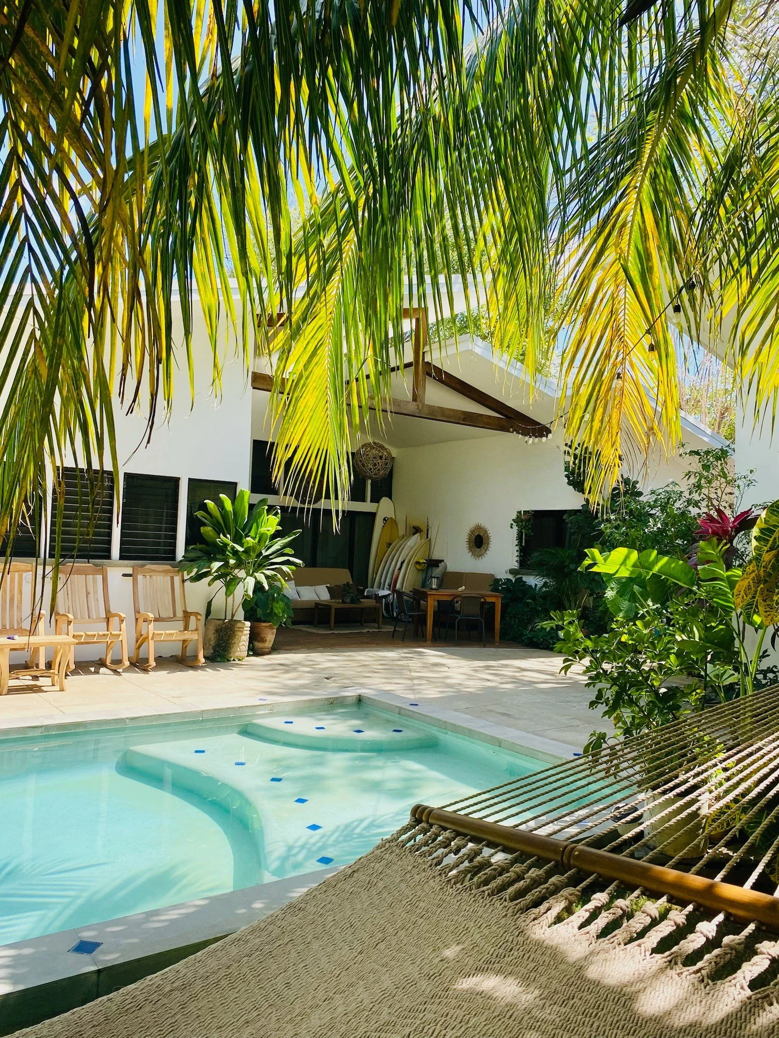 A backyard patio with a small pool, palm trees, potted plants, and outdoor seating under a partially shaded area.