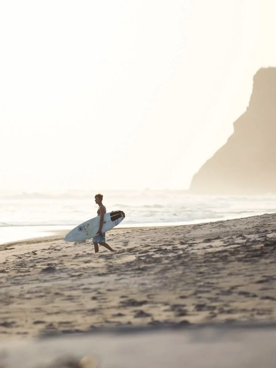 A person walking along the beach carrying a surfboard at sunset or sunrise with a cliff in the background.