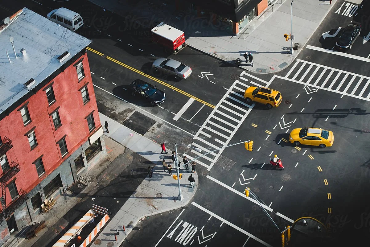 Aerial view of an urban intersection with yellow taxis, cars, pedestrians, crosswalks, traffic lights, and a red brick building on the corner.