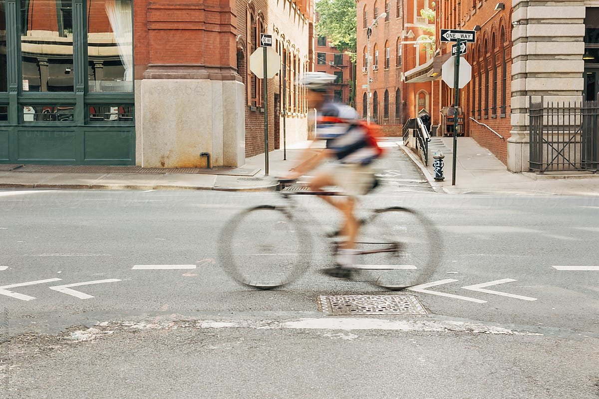 A person riding a bicycle across an intersection in New York City with brick buildings, street signs, and a sidewalk visible.