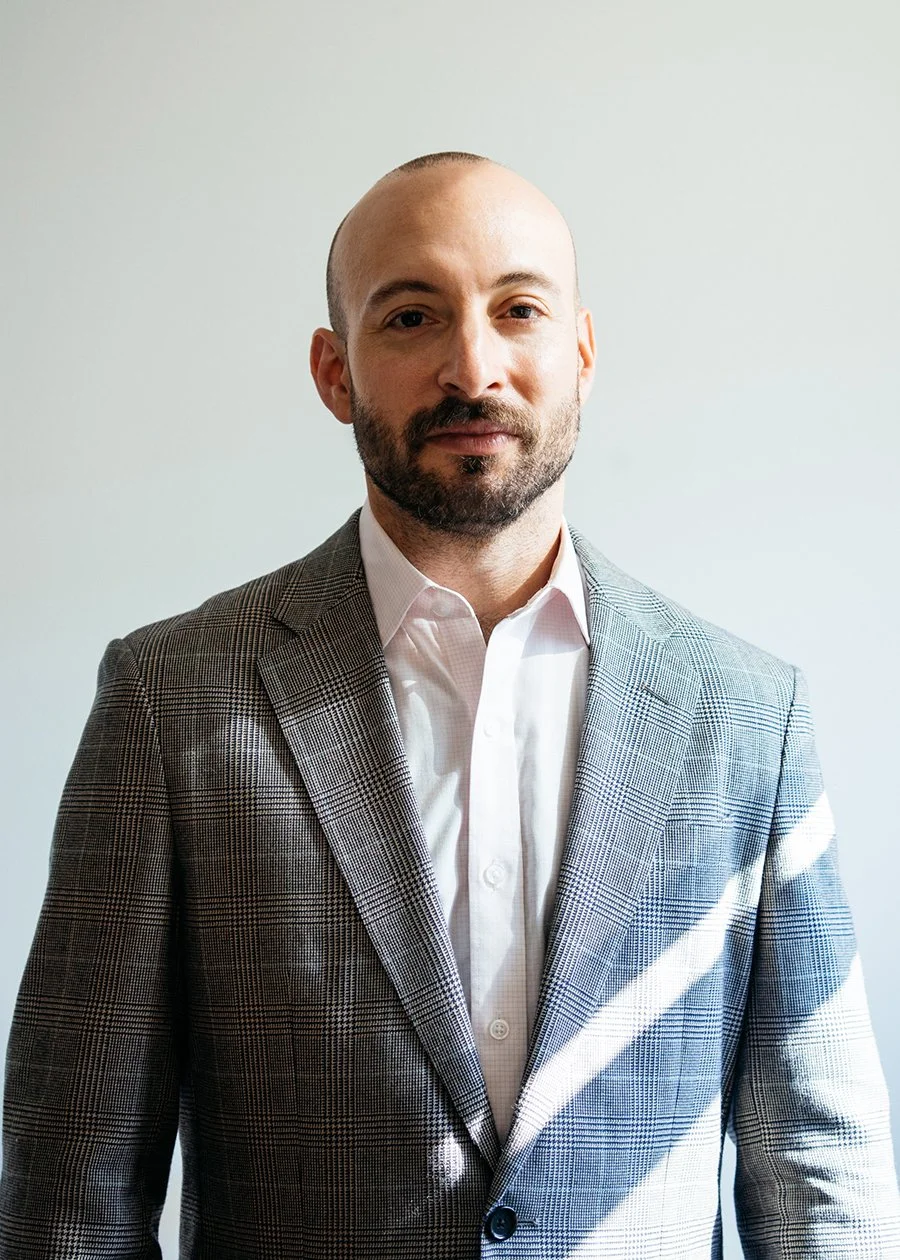 A man with a shaved head and beard wearing a checked blazer and white dress shirt standing against a plain light-colored background.