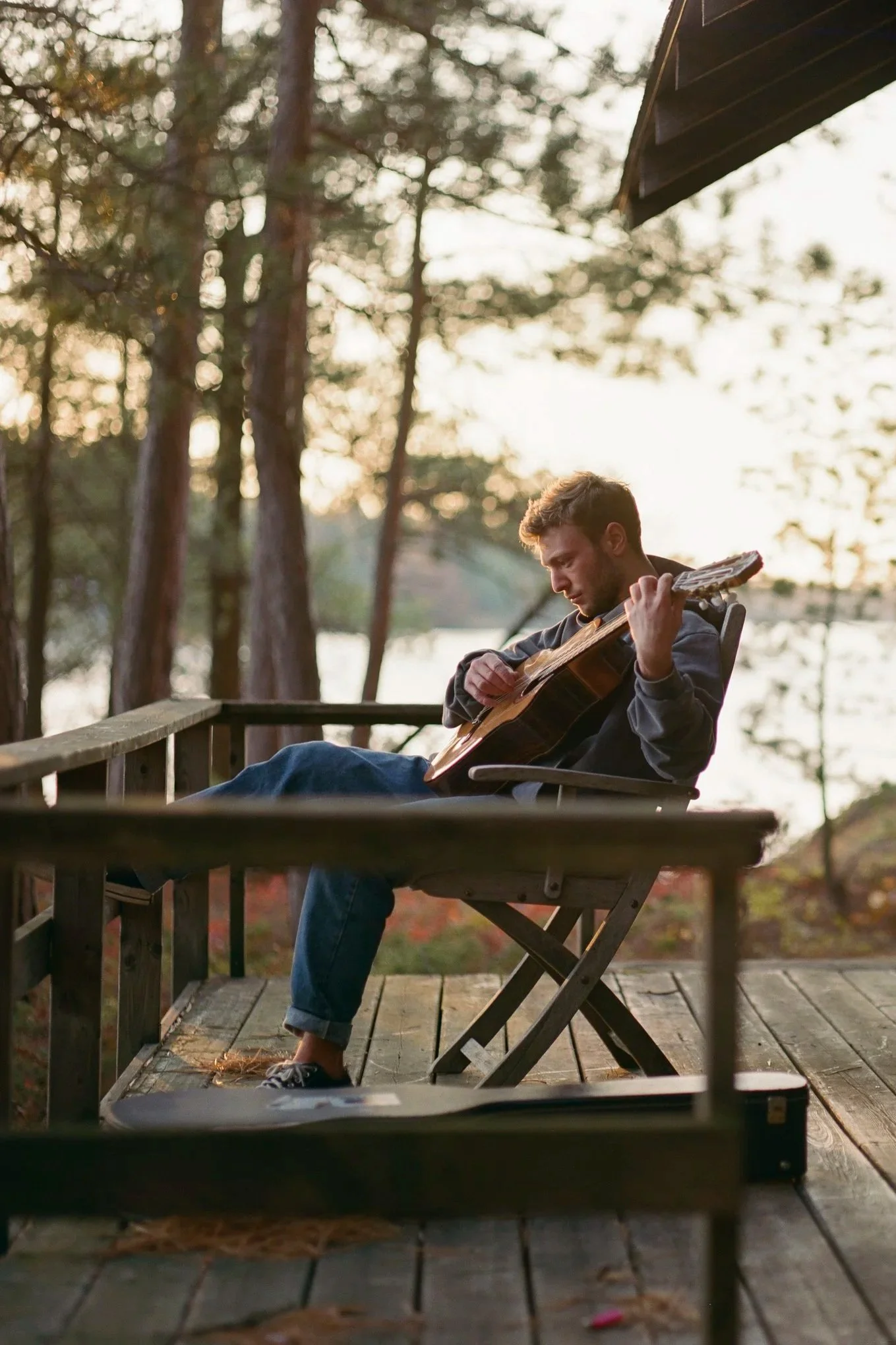 A man sitting on a wooden deck playing an acoustic guitar during sunset in a natural setting with trees and a lake in the background.
