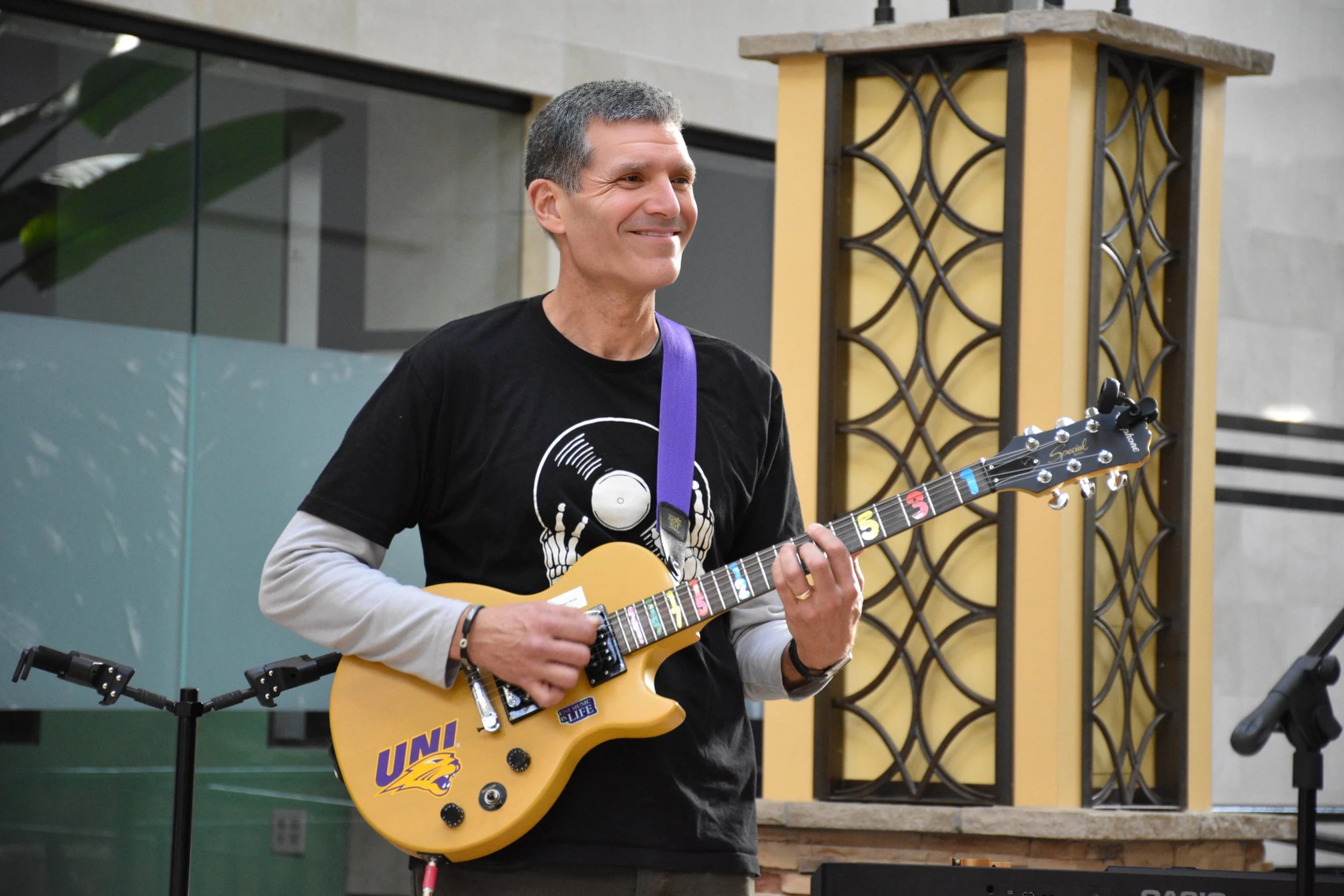 A man is playing an electric guitar in an indoor space, smiling, wearing a black T-shirt with a graphic, with a purple guitar strap. There is a decorative screen behind him and a microphone stand nearby.