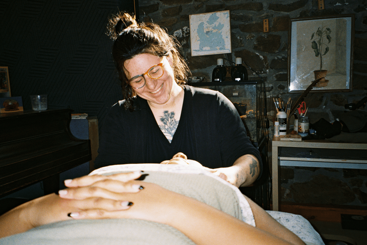 A woman with glasses and dark hair styled in a bun is smiling while giving a tattoo to a person lying down. The background features a stone wall with framed artwork, a piano, and tattoo supplies on a table.