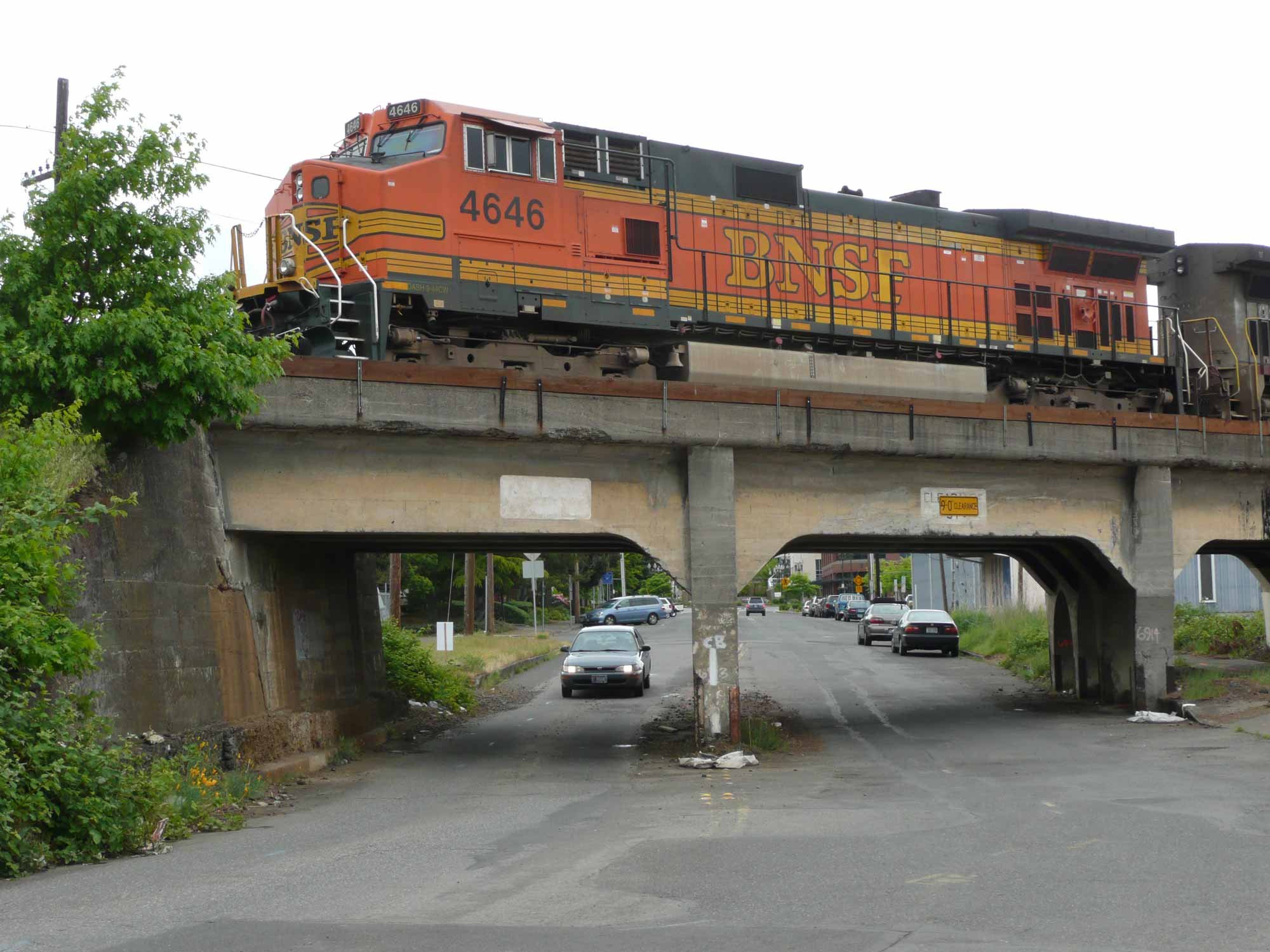 A BNSF freight train engine painted in orange with yellow and black stripes is traveling over a highway bridge, visible from below with several cars passing beneath.