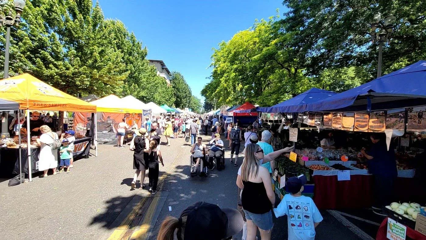 Outdoor farmers market with vendor stalls and shoppers on a sunny day, tall trees lining the street.