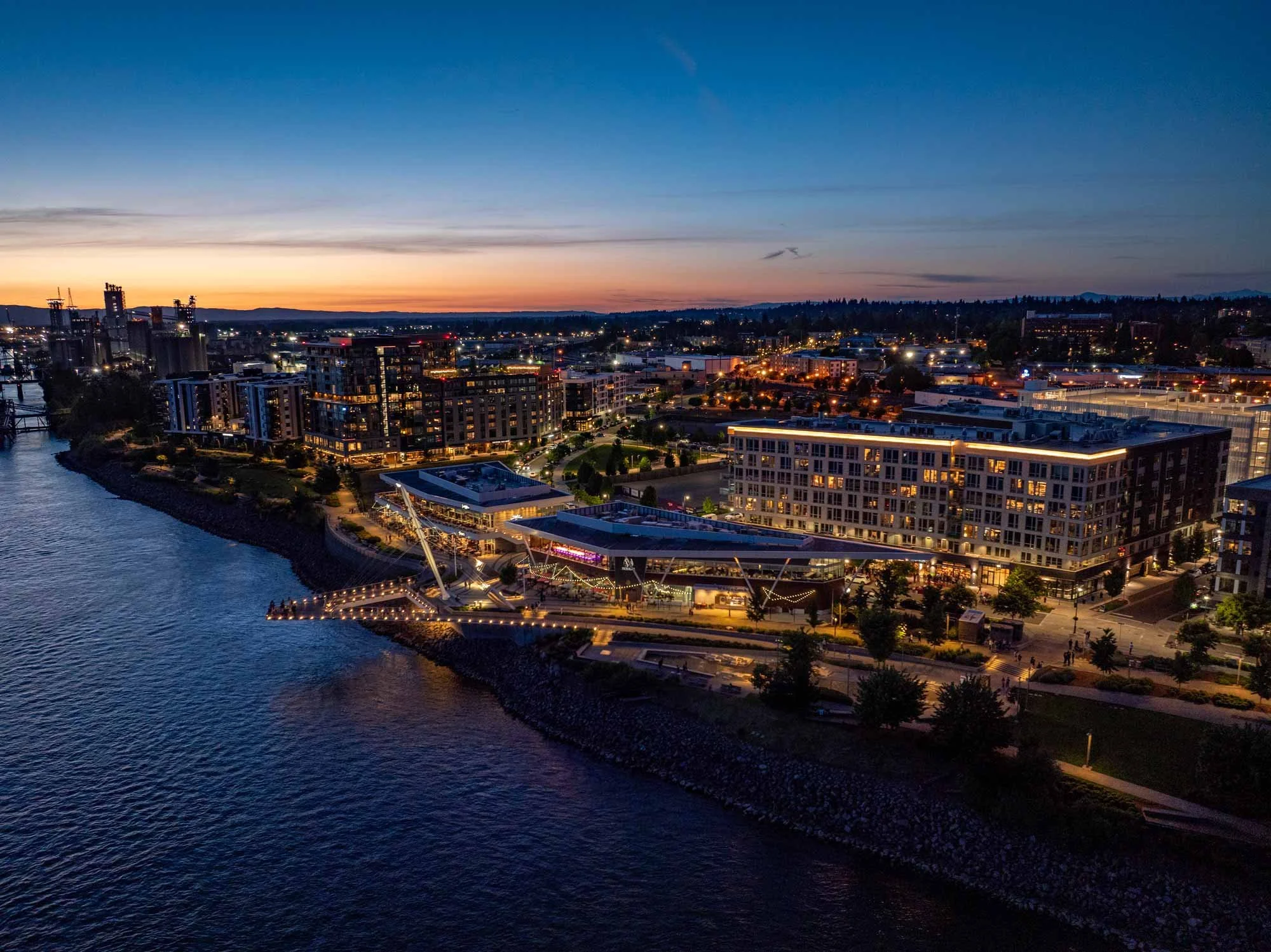 A cityscape at dusk with buildings illuminated and a river in the foreground. The sky shows a gradient from orange to dark blue.