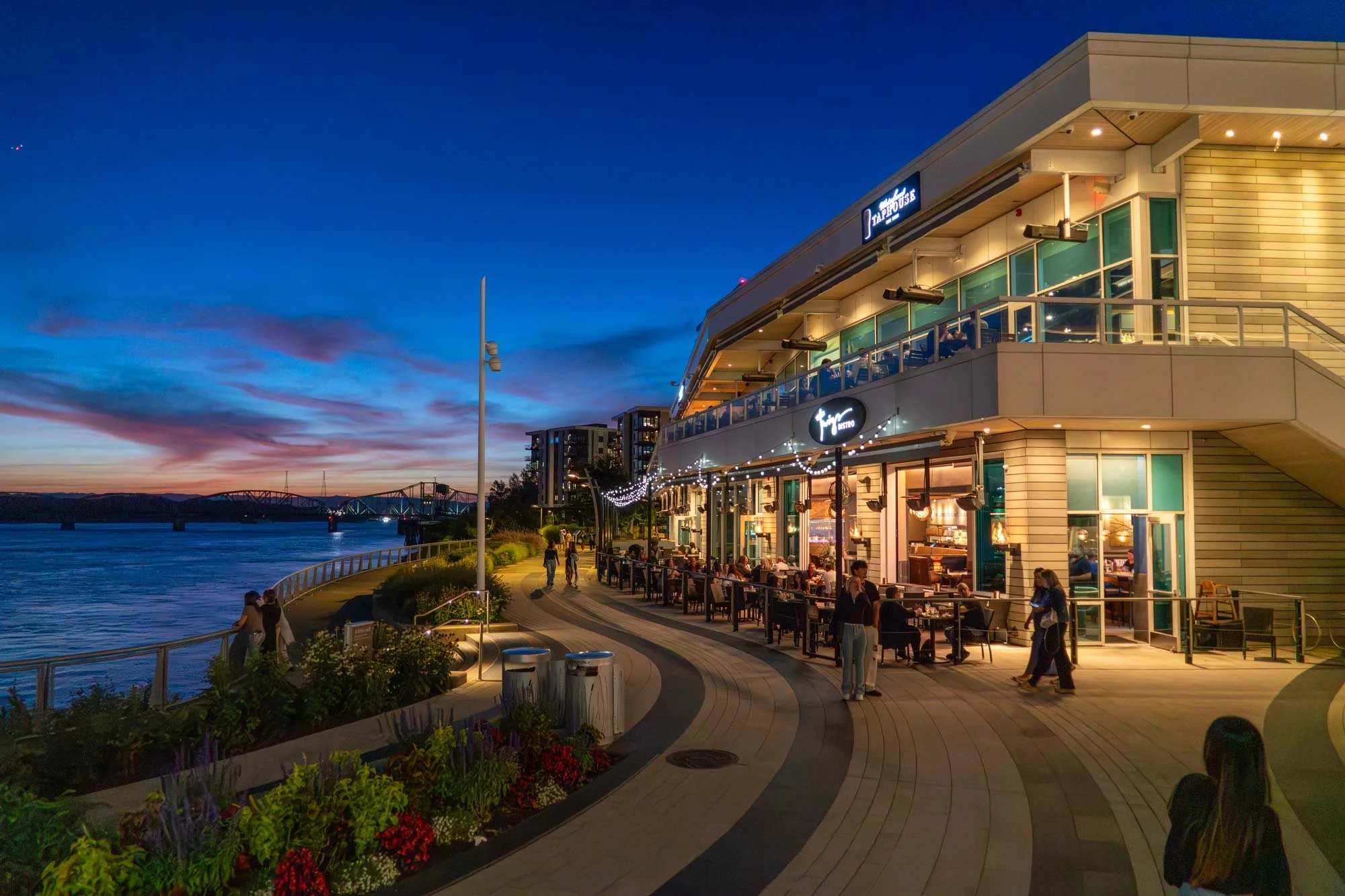 Exterior of a waterfront restaurant or café during the evening with people dining outside under string lights, overlooking a river or bay with a bridge in the distance and colorful sunset sky.