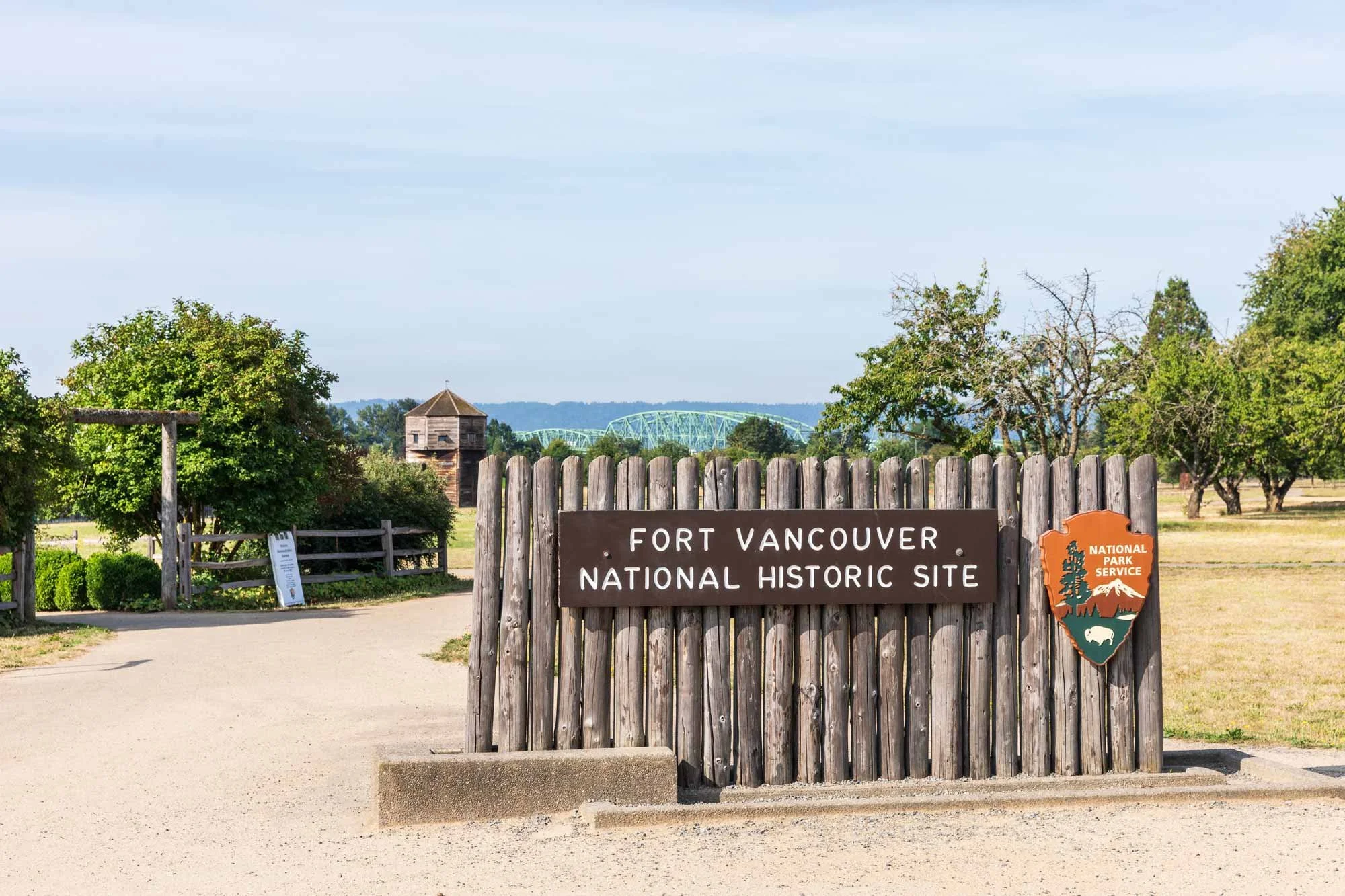 Entrance sign for Fort Vancouver National Historic Site, with wooden fence, trees, and a historical building in the background.