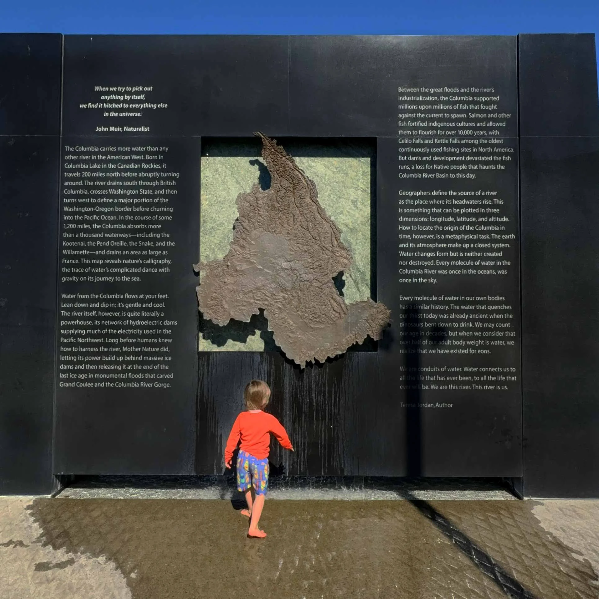 A boy wearing a red long-sleeved shirt and colorful shorts stands barefoot on wet ground, facing a large black informational display about the Columbia River, with a small sample of rock or earth in front of it.