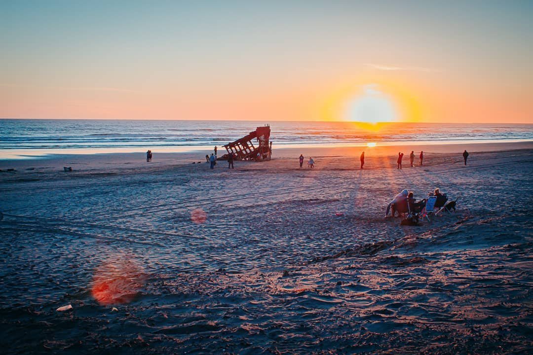Sunset over the ocean with a sandy beach, people enjoying the scenery and others sitting on chairs, a partially submerged wooden structure, and the horizon in the background.