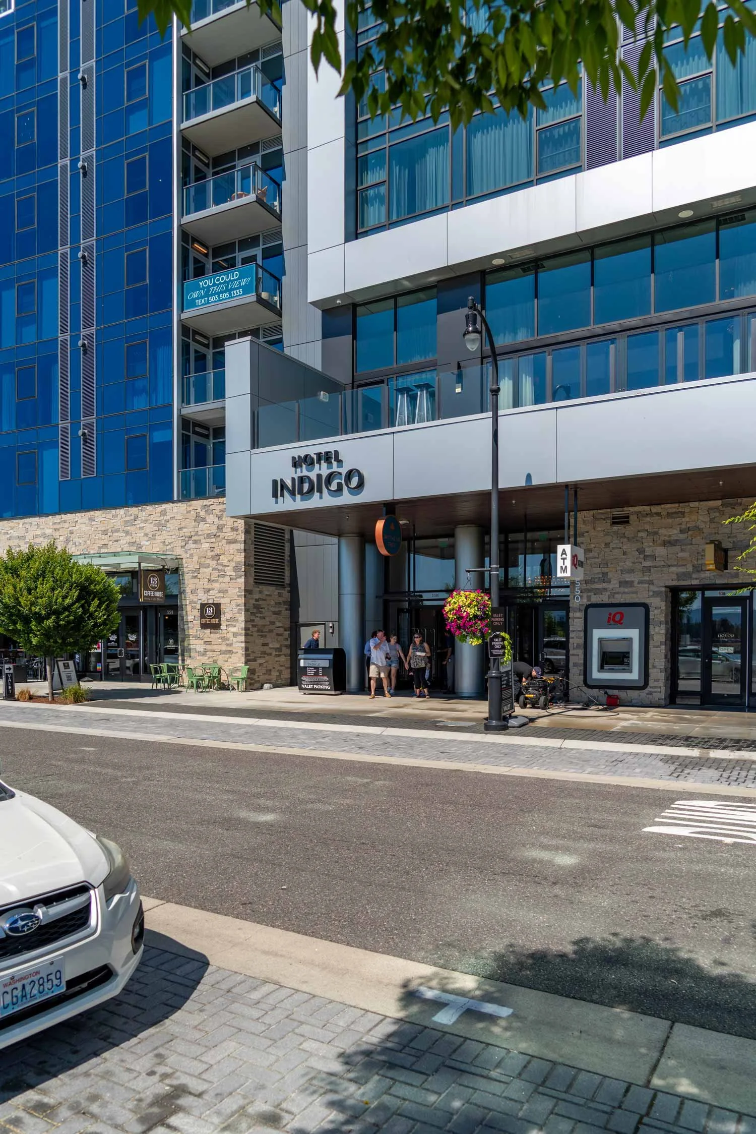 A street view of Hotel Indigo, a modern building with glass windows and balconies. There are people walking near the entrance, parked cars, a streetlamp, and a parking meter.