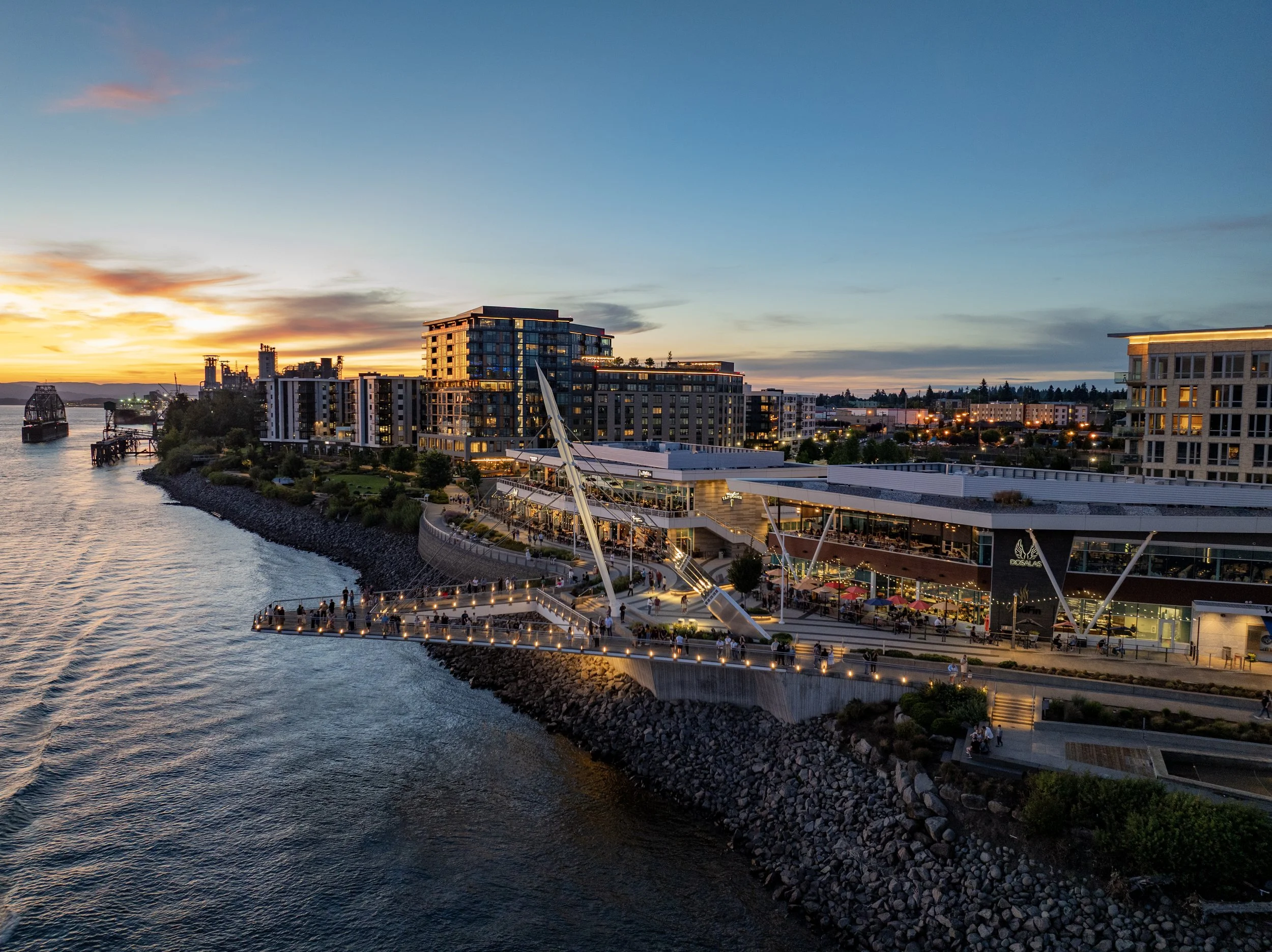 A city waterfront at sunset with high-rise buildings, a promenade filled with people, and a body of water with boats.