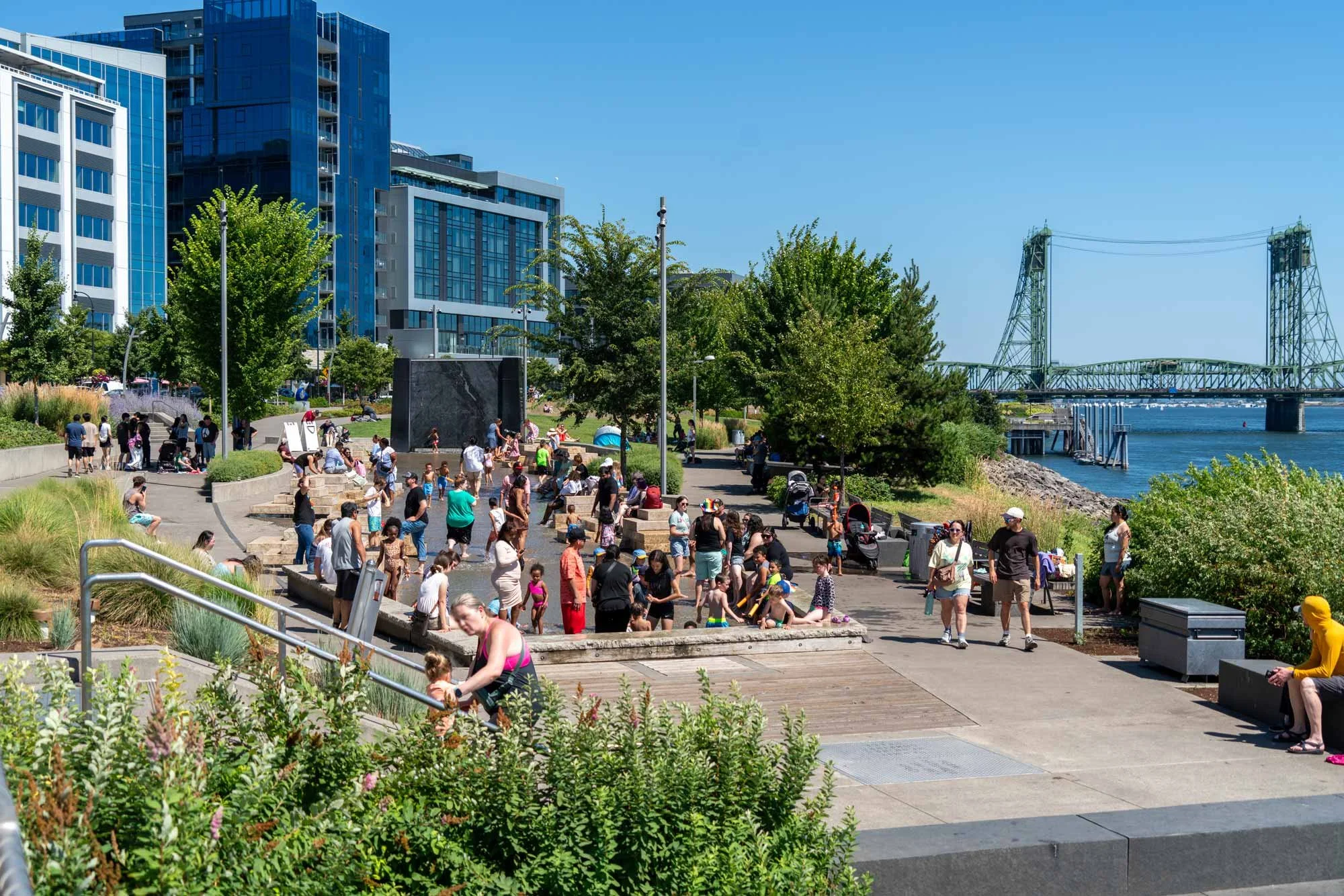 A busy waterfront park with many people walking, sitting, and enjoying the sunny day. There are tall modern buildings, green trees, and a bridge over the water in the background.
