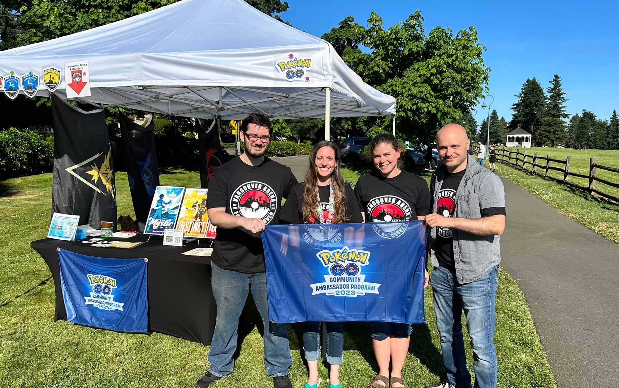 Four people standing under a white canopy tent with Pokémon GO banners, holding a large blue flag that reads 'Pokémon GO Community Ambassador Program 2023.' They are outdoors on a sunny day with a park and trees in the background.