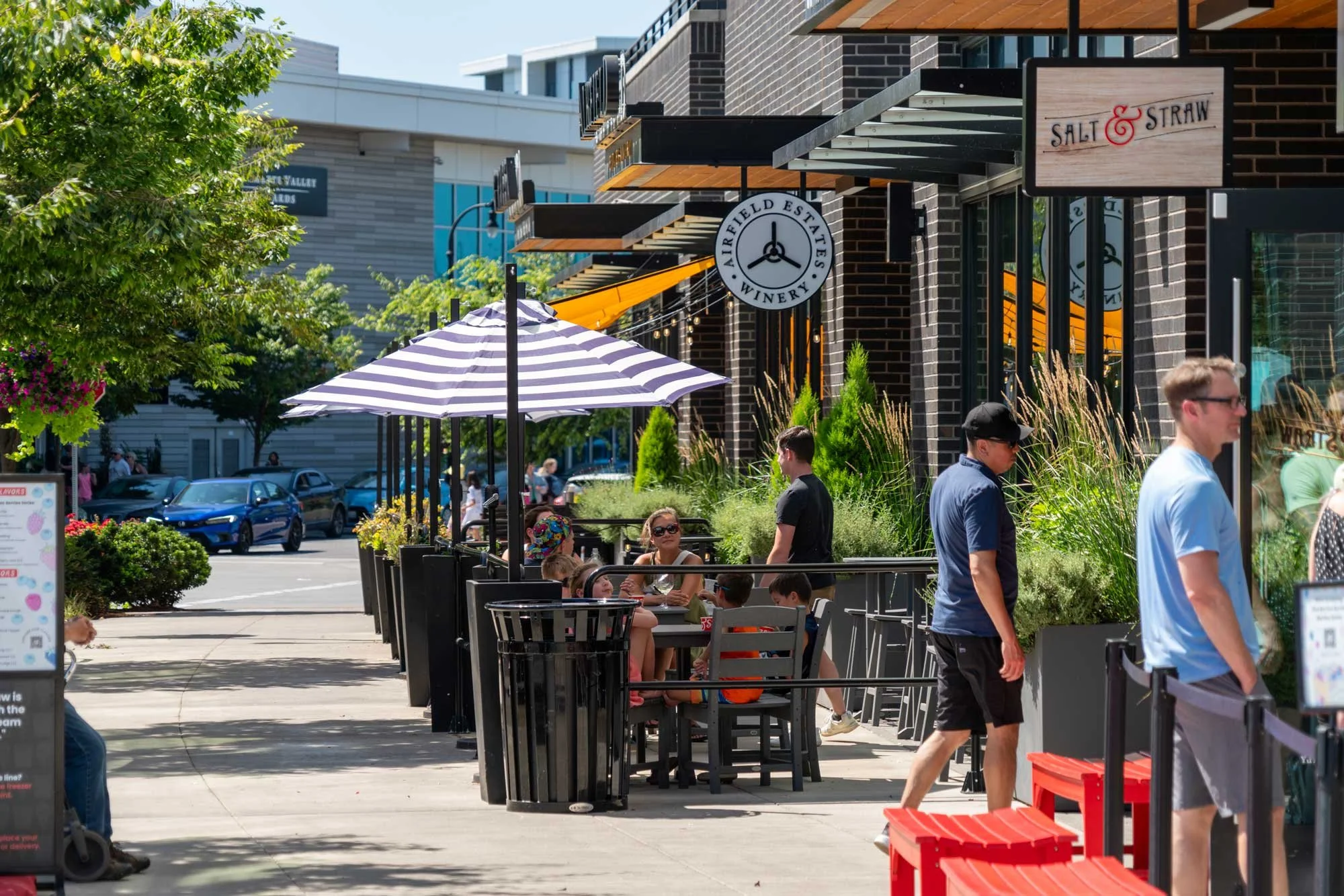 People sitting and eating outdoors at a cafe on a sunny day, with a sign for Salt & Straw ice cream and a winery in the background, under umbrellas and surrounded by greenery.