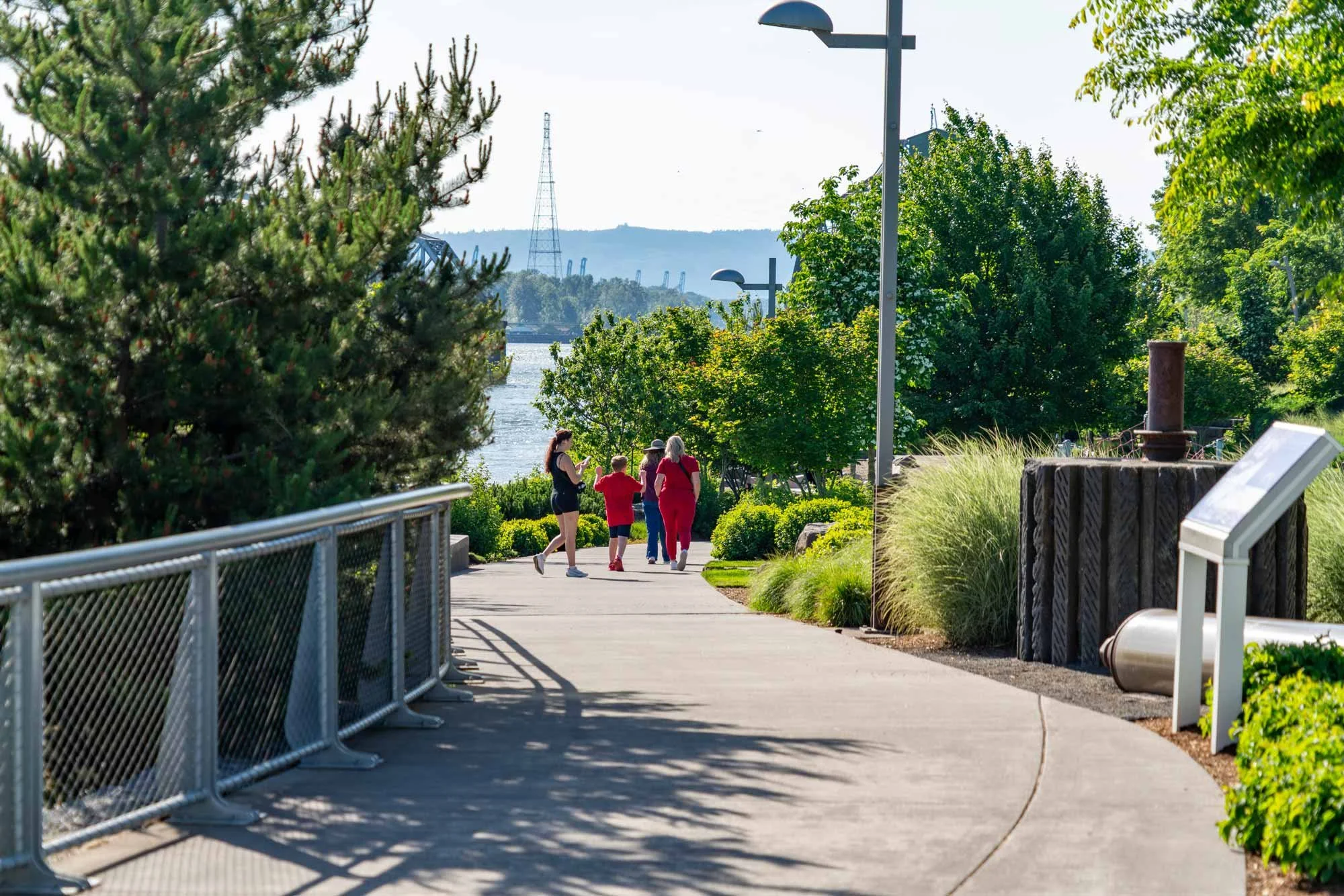 A paved walkway in a park with a group of four people, two adults and two children, walking and talking among lush green trees. A metal railing lines one side of the path, and there are streetlights along the path. In the background, water and industrial structures such as cranes and a tower are visible under a clear sky.