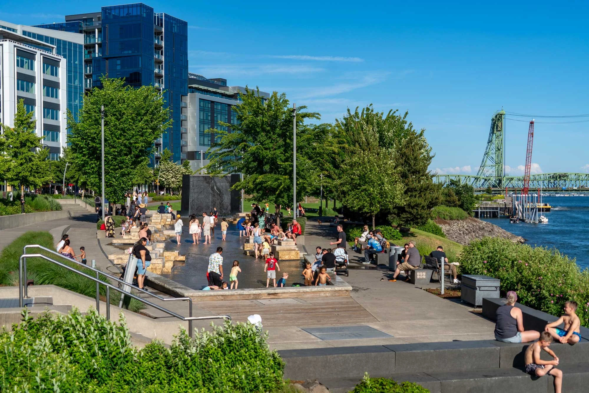 People enjoying a sunny day at a waterfront park with a fountain and trees, surrounded by modern buildings and a bridge in the background.