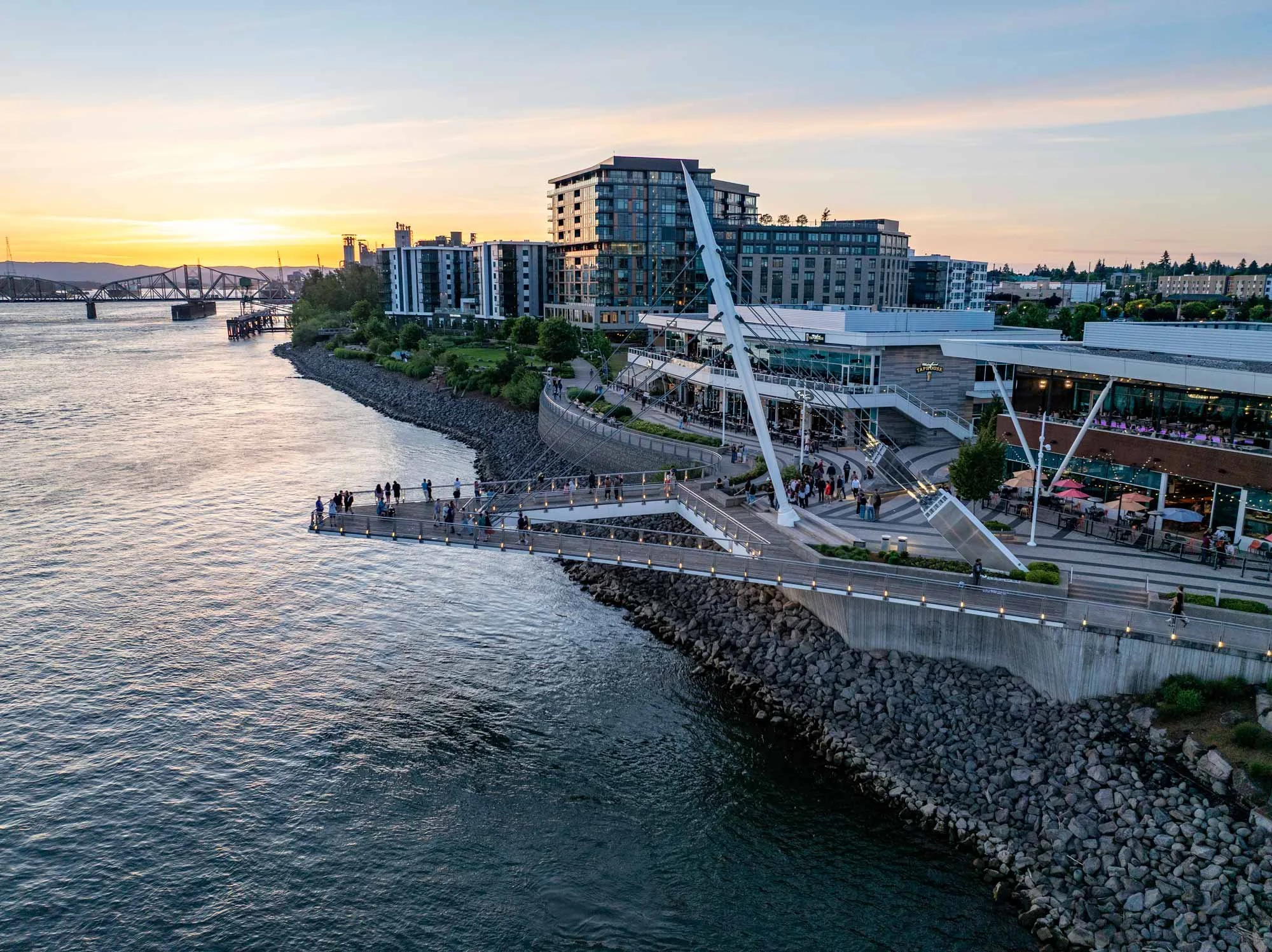 A modern waterfront cityscape during sunset with a river, a pier, high-rise buildings, and a bridge in the distance.