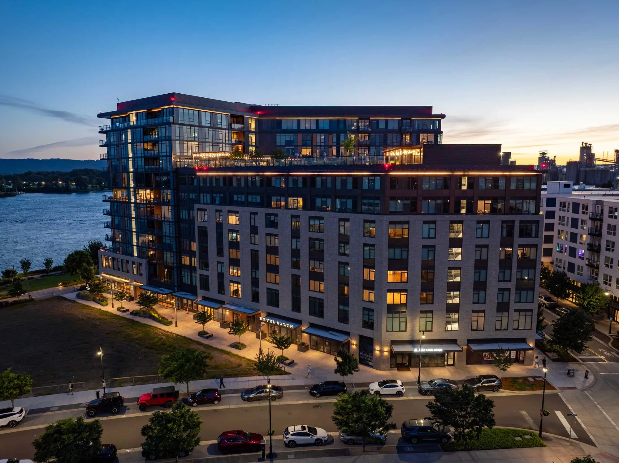 A modern multi-story building illuminated at dusk with glass windows, a rooftop terrace, and a street with parked cars in the foreground.