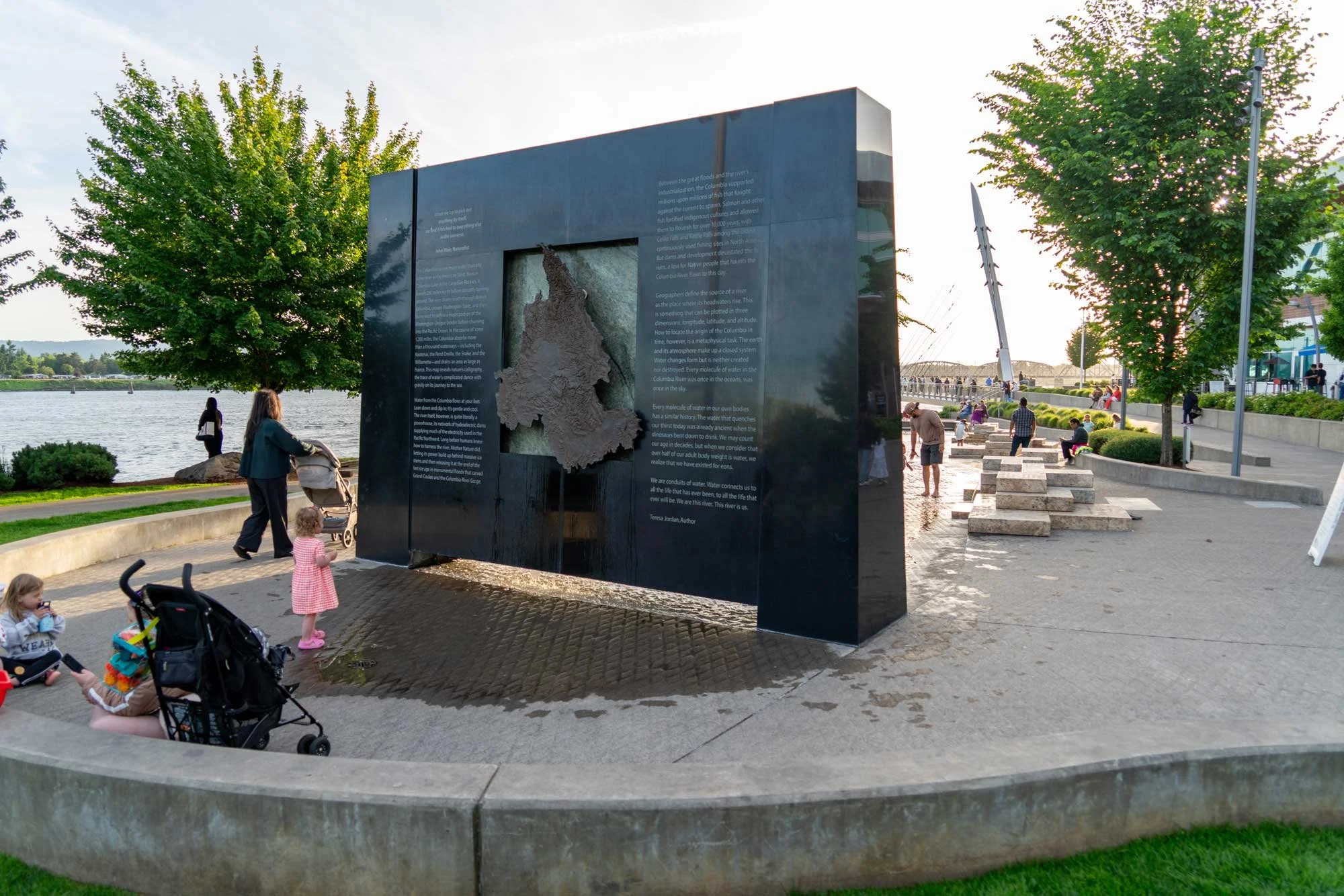 People gather around an informational display about the Columbia River near a waterfront area with trees, benches, and a bridge in the background.