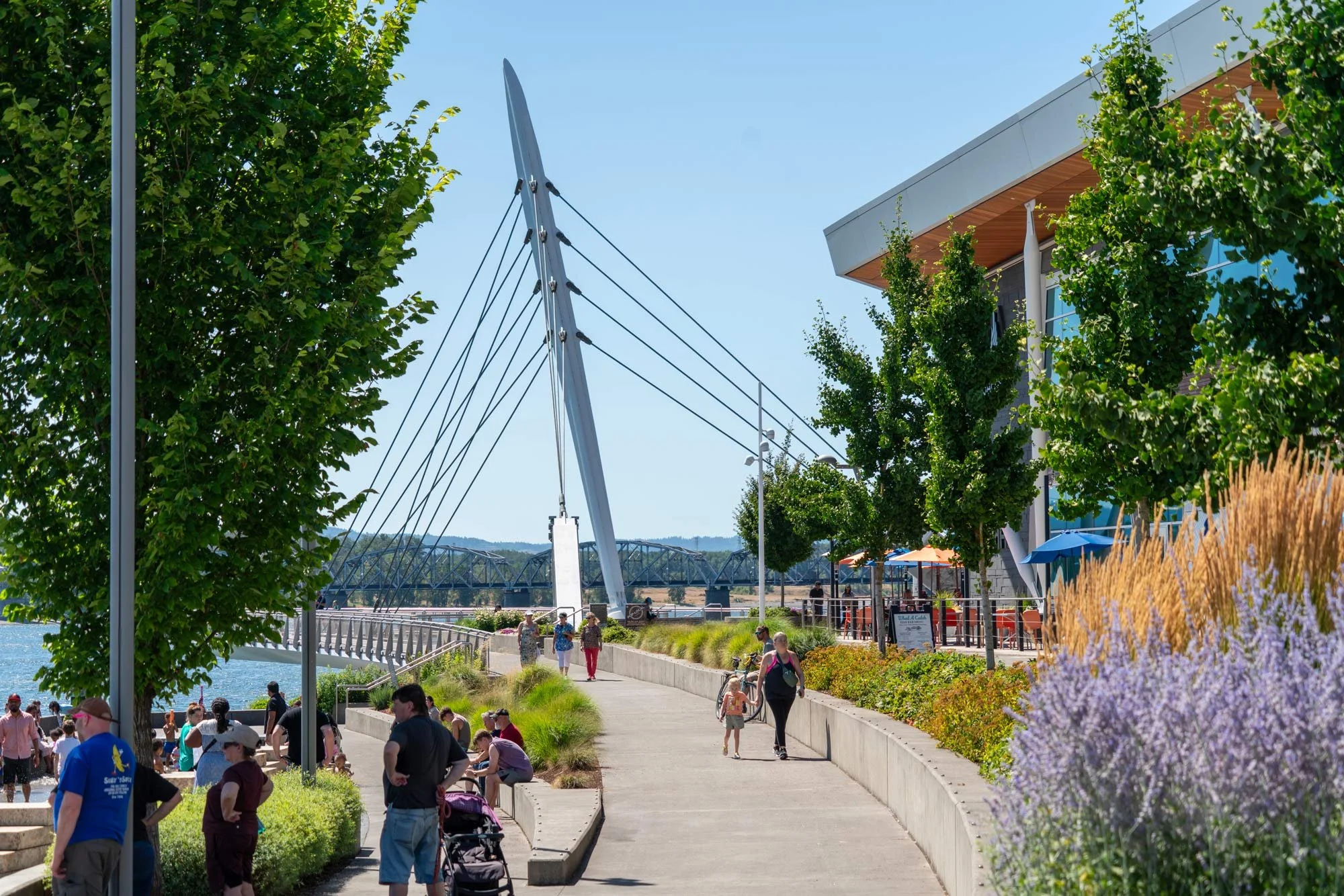 People walking and relaxing in a park along a waterway on a sunny day. There are trees, flowers, a modern sculpture, and a building with outdoor seating.