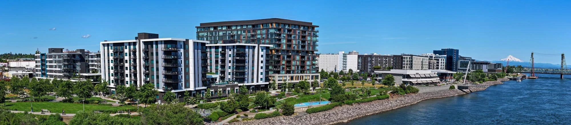 View of modern high-rise residential buildings along a waterfront with a park, walking paths, and lush greenery, with a bridge and mountain in the background.