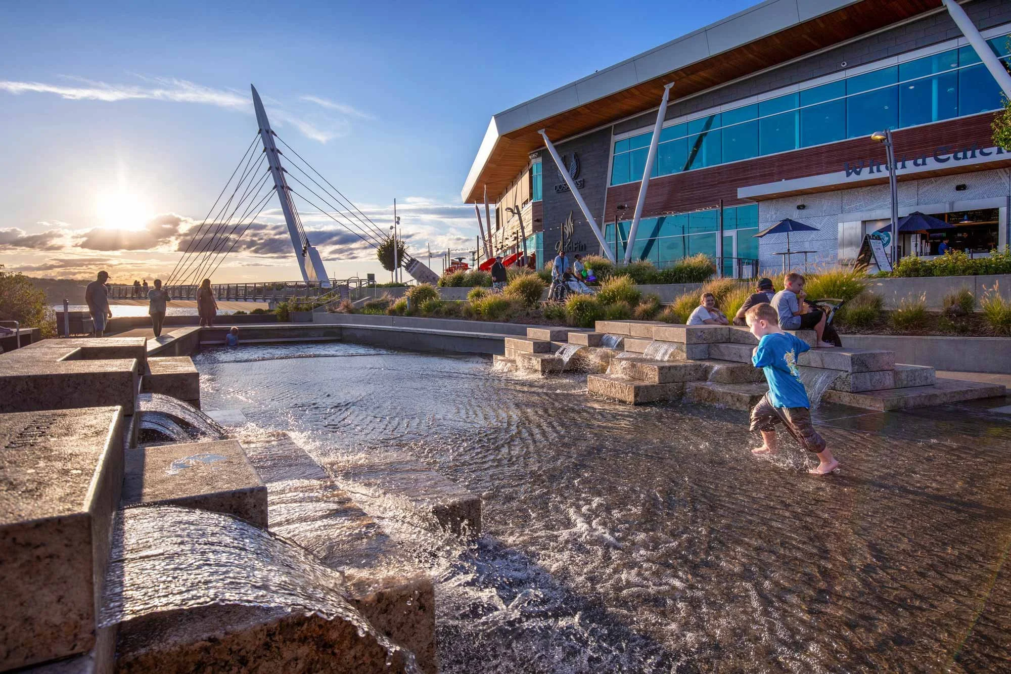 People enjoying a small fountain at sunset near a modern building with large glass windows.
