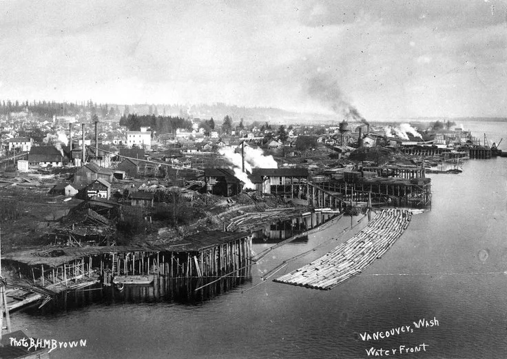 Historical black and white photograph of the waterfront in Vancouver, Washington, showing industrial buildings and smoke in the background, with a wooden dock extending into the water.