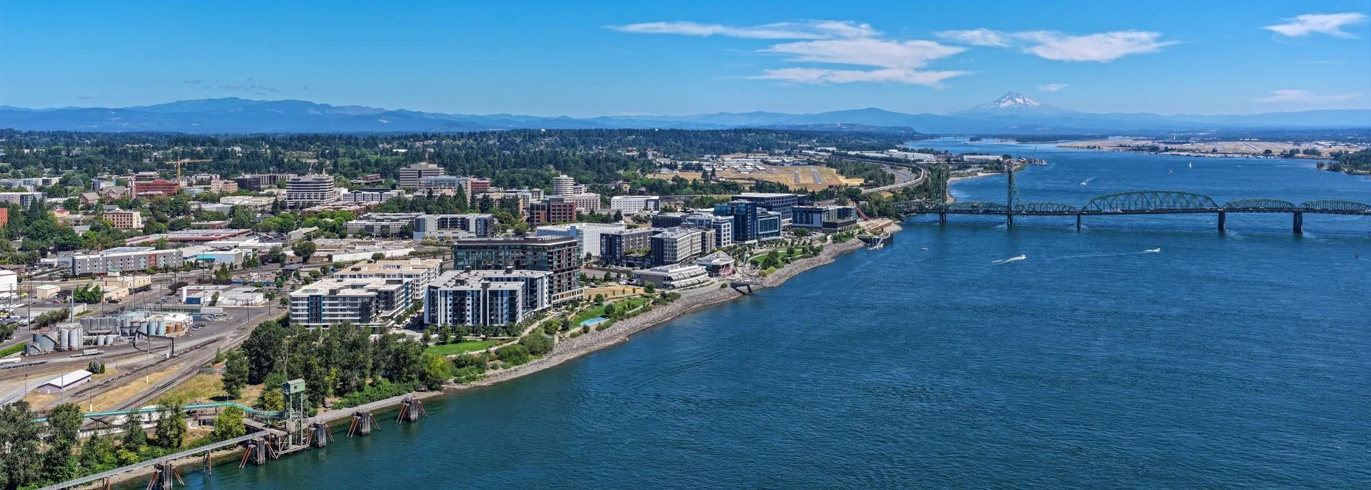 Aerial view of a city along a river with modern buildings, a bridge, and distant mountains including Mount St. Helens.