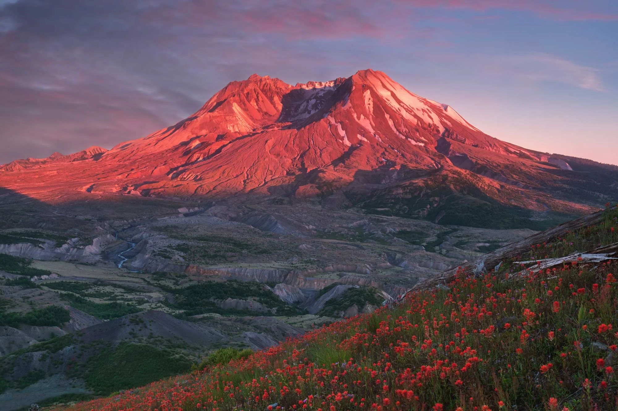 A scenic mountain landscape at sunset, featuring a tall rocky mountain with snow patches and a reddish glow, surrounded by green hills, rocky terrain, and bright red flowers in the foreground.