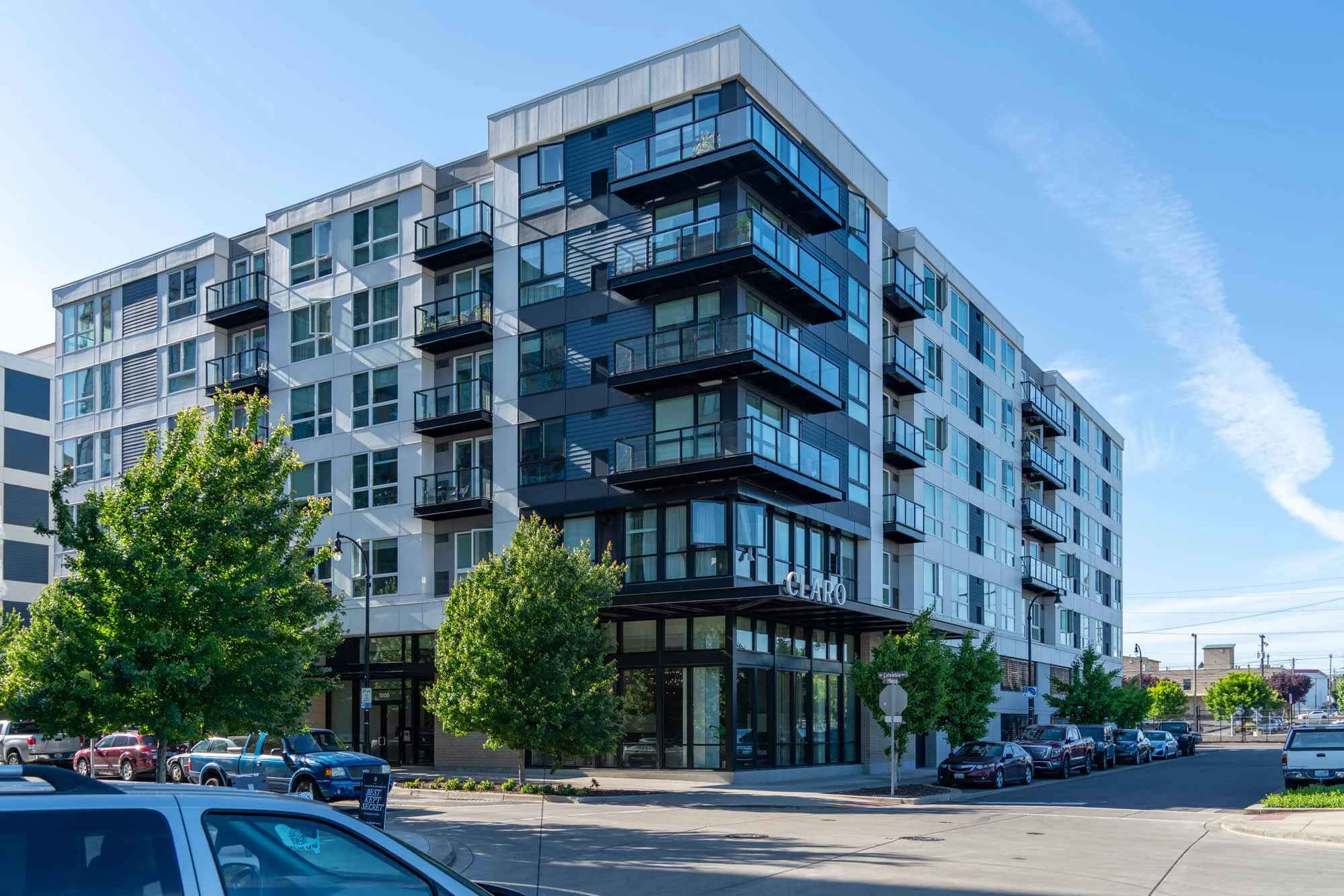 Modern multi-story residential building with glass balconies and a mix of white and dark exterior siding, surrounded by green trees and a parking lot with cars, under a blue sky with some clouds.
